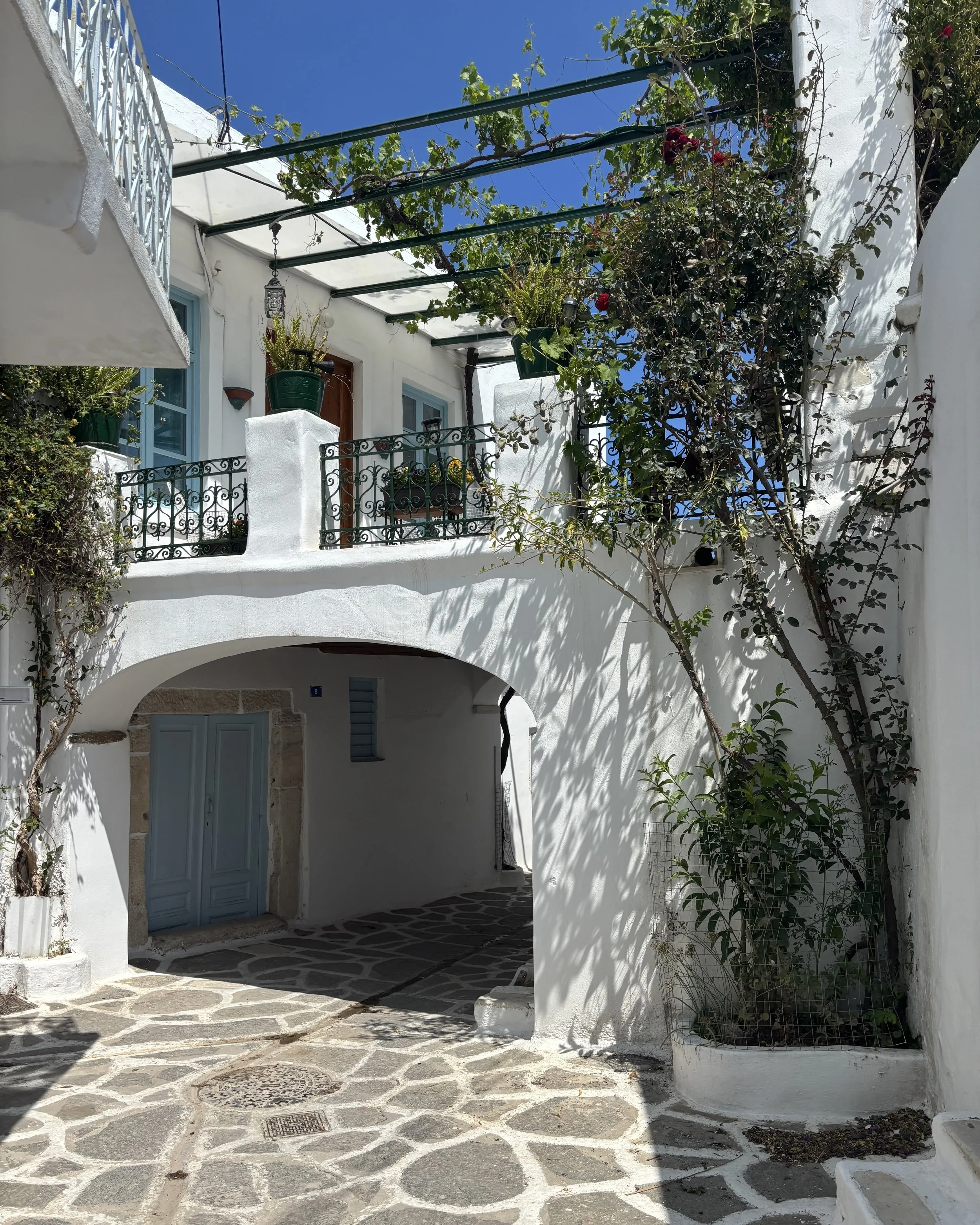 White Mediterranean-style building with blue window frames, a small arched passageway, and a cobblestone pathway, with climbing plants and a balcony with potted plants under a clear blue sky.