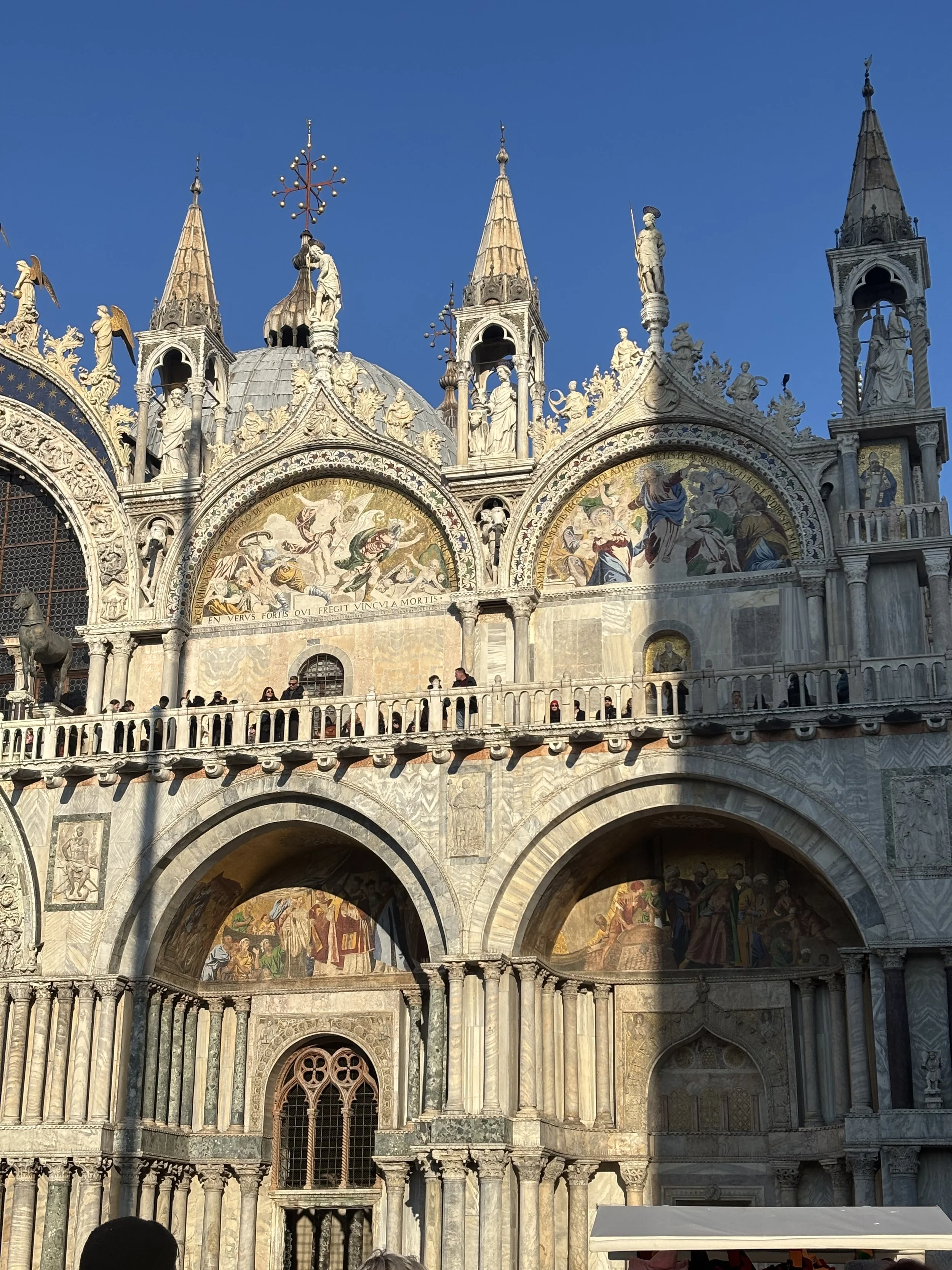 Front facade of St. Mark's Basilica in Venice, Italy, showing ornate architecture, mosaics, statues, and turrets against a blue sky.
