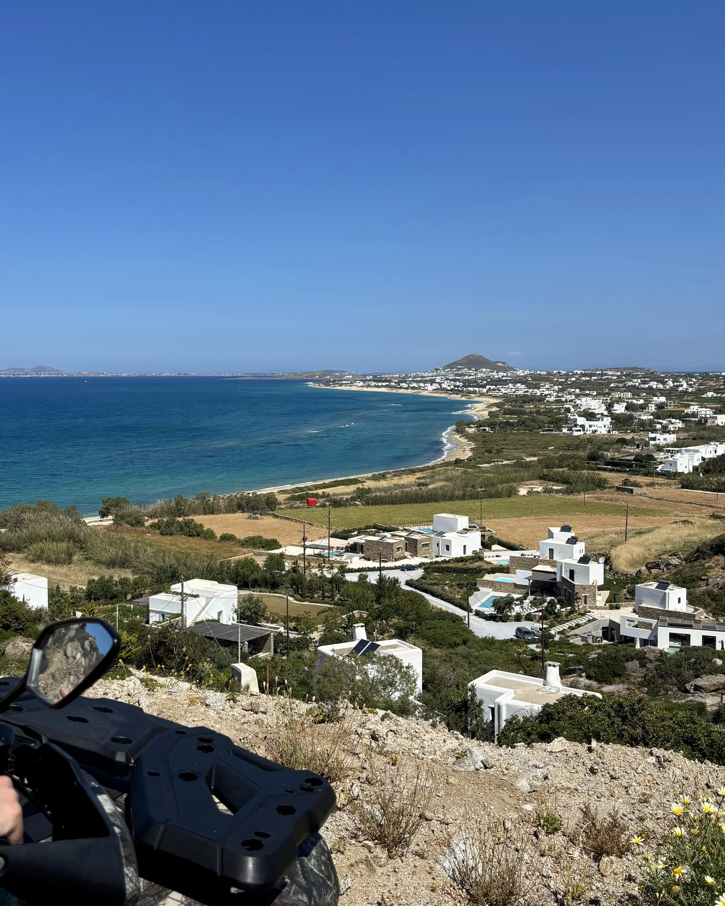 A coastal landscape with a blue ocean, sandy beach, white modern houses, and distant hills on a sunny day with a motorcycle in the foreground.