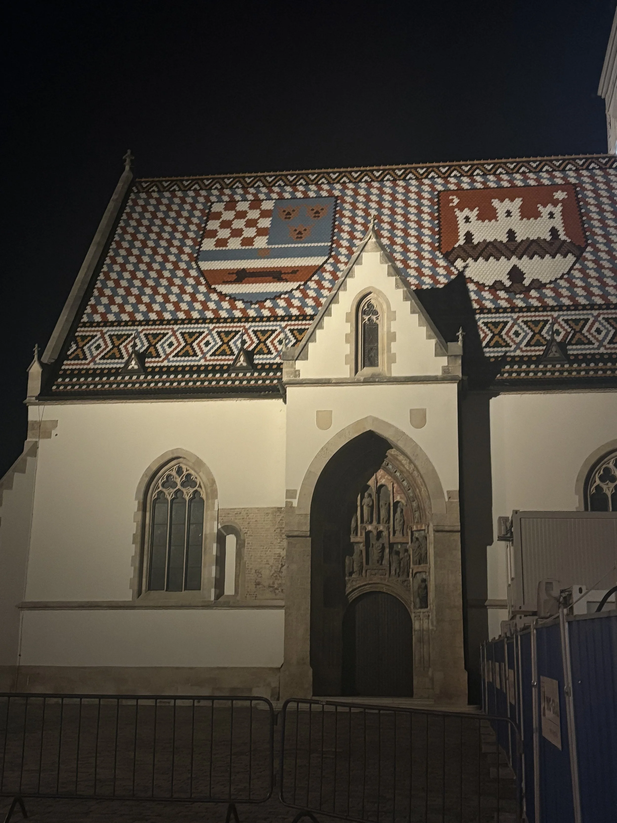 Night view of a historic church with a colorful patterned tiled roof featuring two heraldic shields. The church has Gothic-style arched windows and an ornate arched doorway with sculptures and carvings, illuminated by external lights at night.