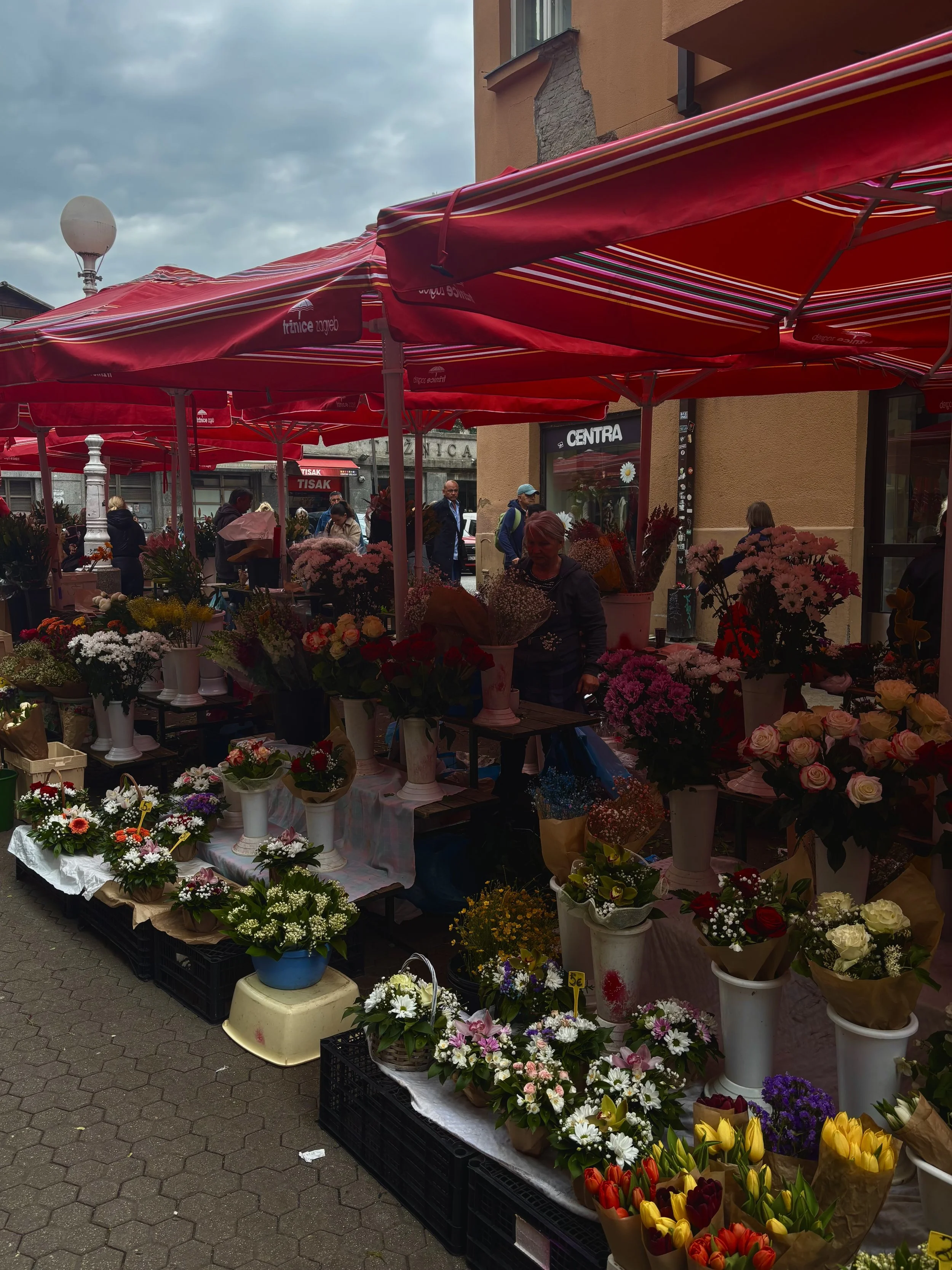 An outdoor flower market with various colorful flowers in vases and baskets under red umbrellas.