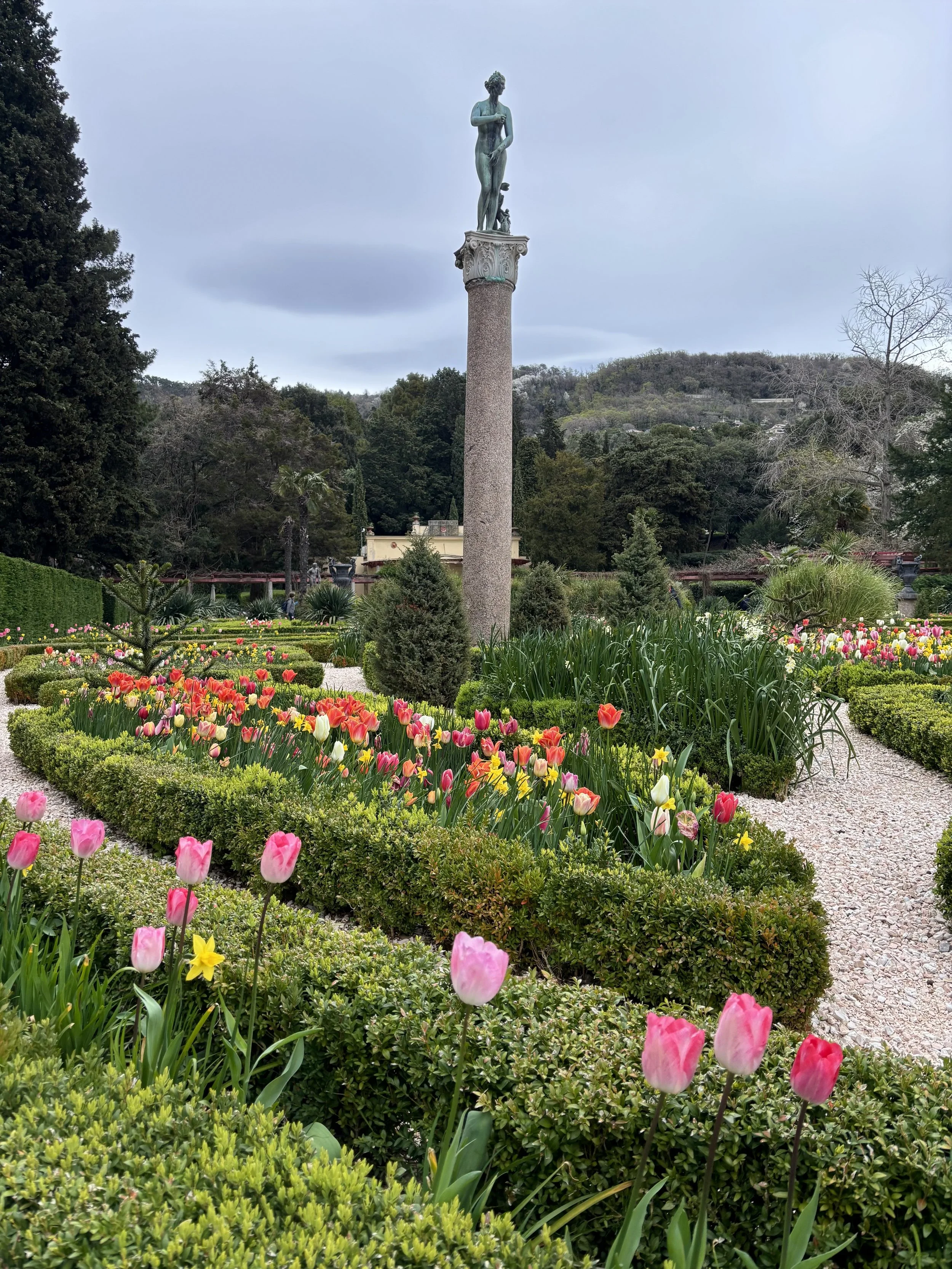 A garden with colorful tulips and yellow flowers arranged in symmetrical beds, with a tall column topped by a statue of a standing woman, surrounded by lush green trees and hills in the background.