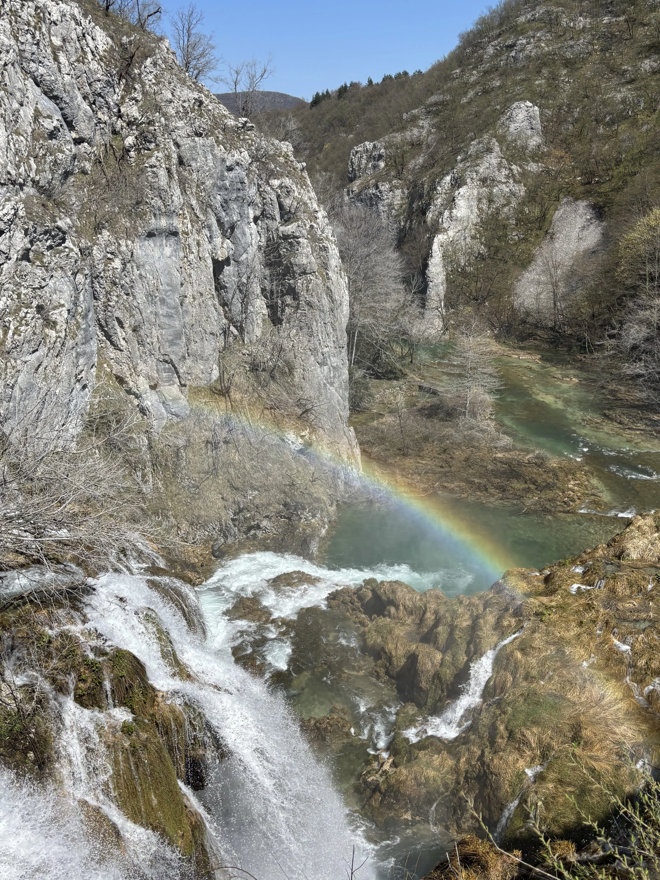 A scenic view of a river cascading over rocks in a canyon with a rainbow arcing across the waterfall and cliffs in the background.