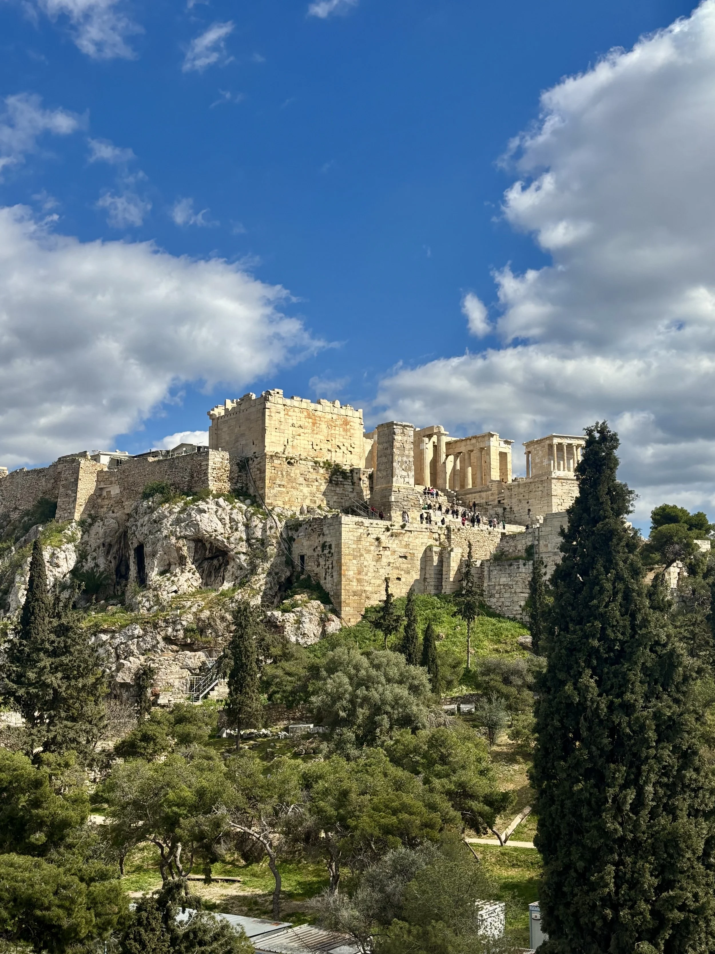 Ancient ruins of the Acropolis in Athens, Greece, surrounded by trees under a partly cloudy sky.