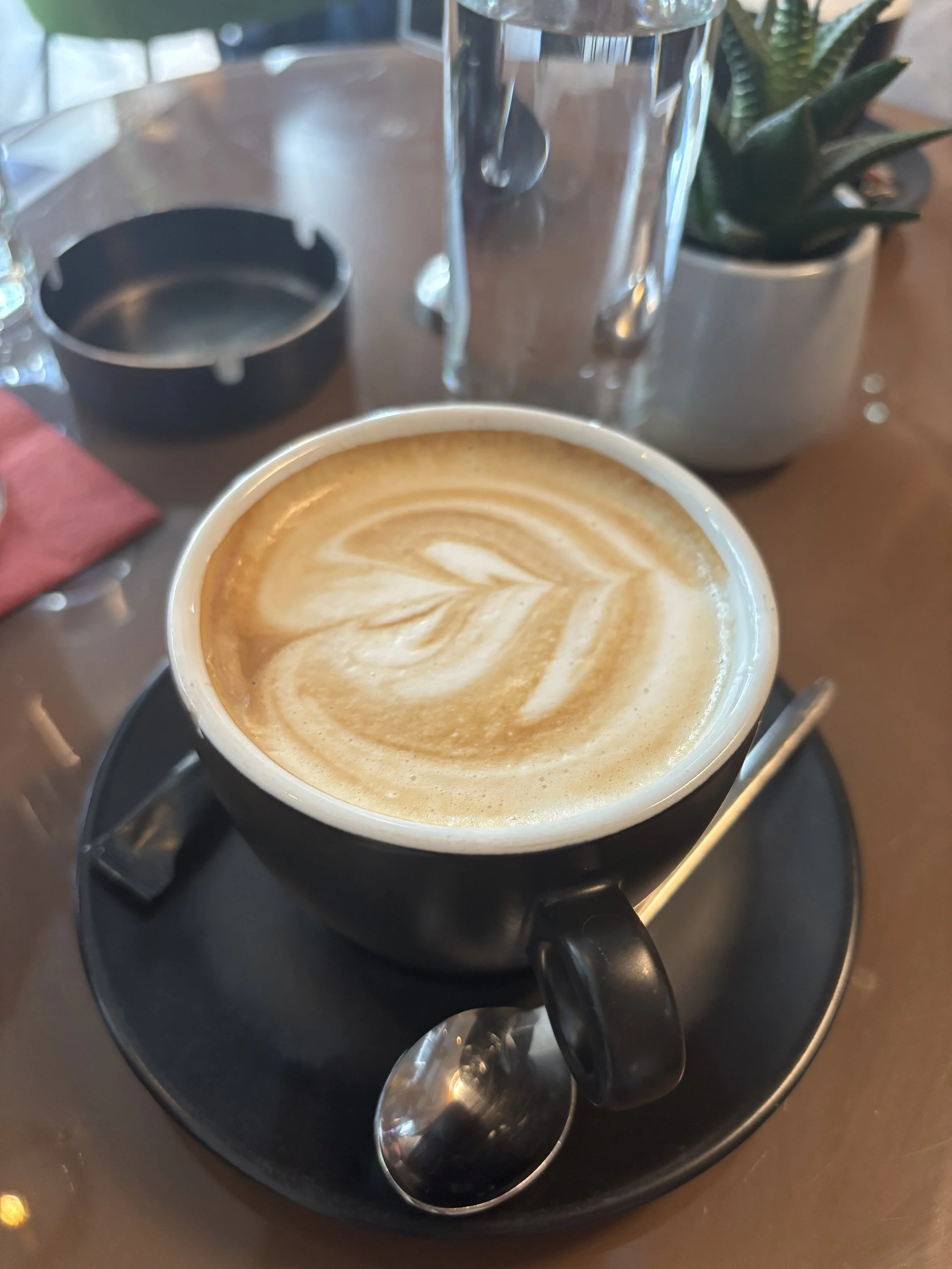 A cup of coffee with latte art on a black saucer, spoon, and small wrapped sugar on a wooden table, with a plant and glass of water in the background.