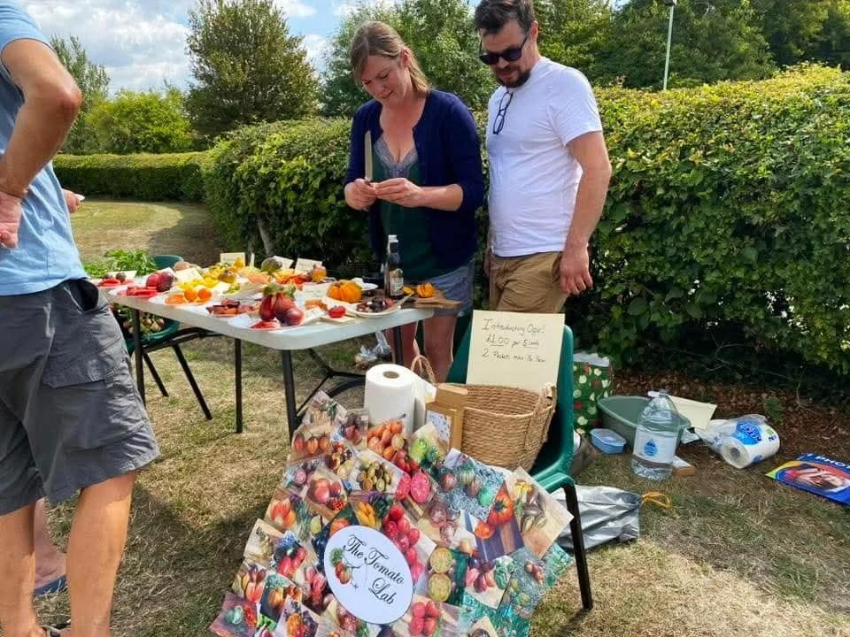 People at a fall-themed outdoor event selling decorative painted eggs and pumpkin crafts. A woman and man are behind a table filled with colorful painted eggs, pumpkins, and other items. A sign on the table reads, "Introducing Our $2 Egg".