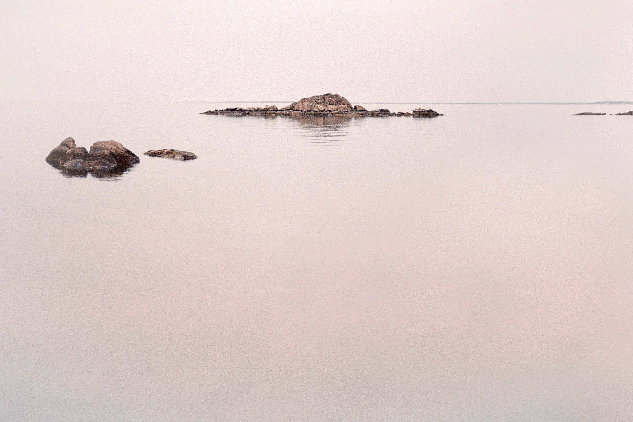 A calm body of water with several rocks protruding above the surface, and a small rocky island in the distance, under a pale, overcast sky.