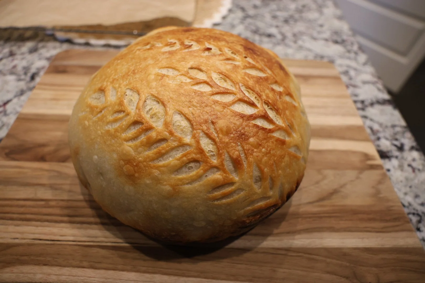 A round loaf of bread with decorative slits on top, sitting on a wooden cutting board