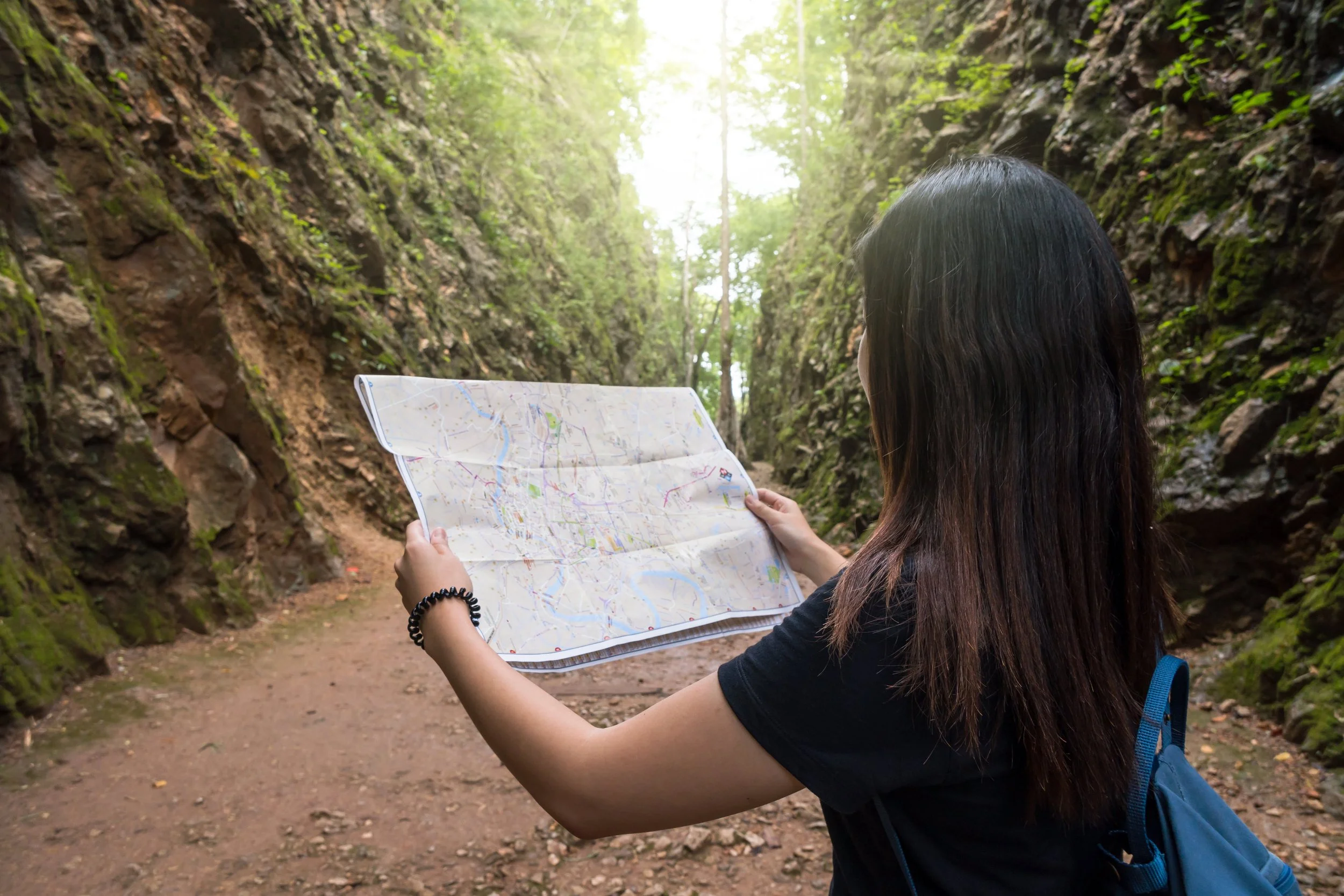A person with long dark hair holding a map in a narrow, rocky canyon surrounded by green foliage.