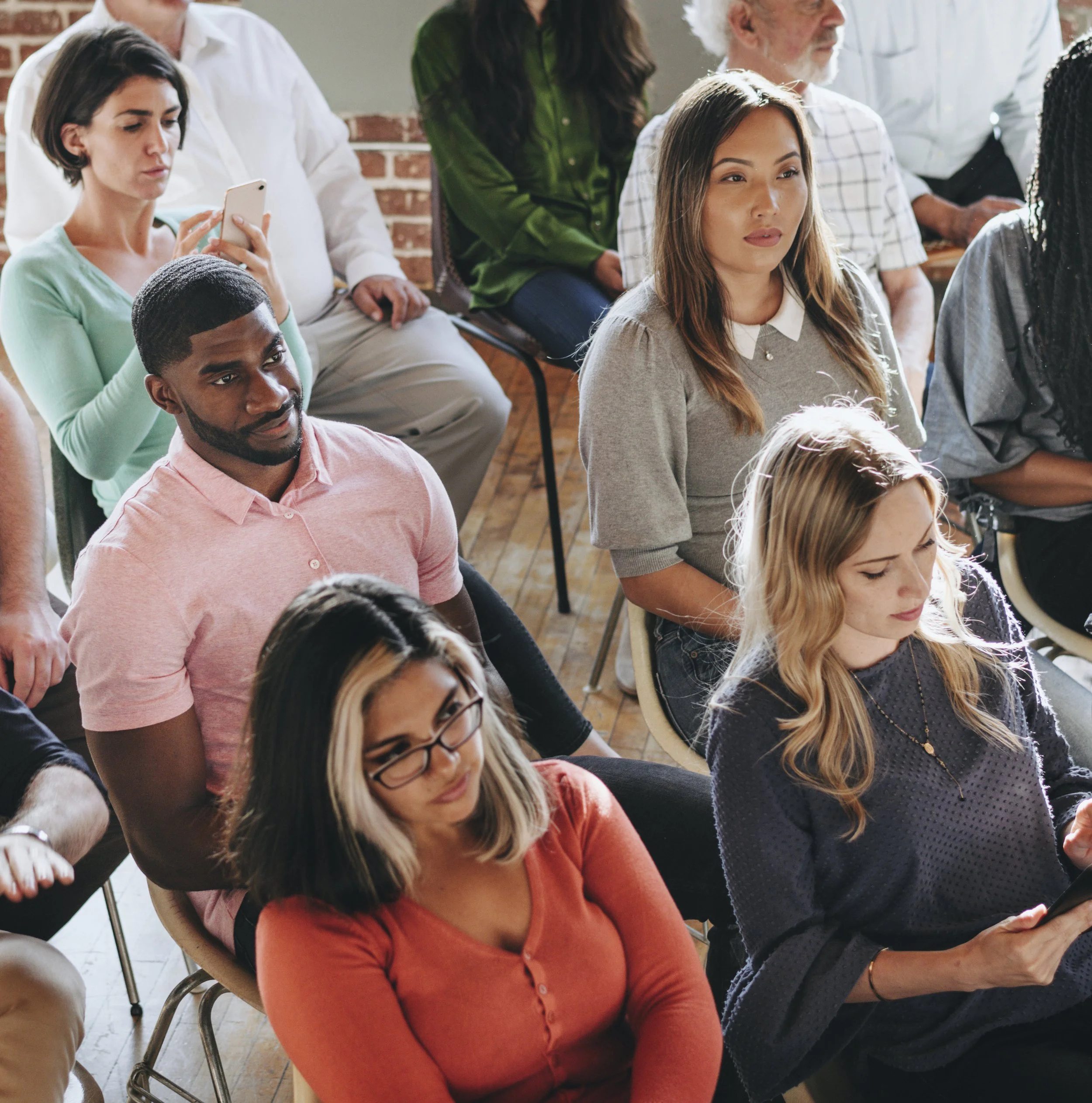 A group of people sitting and listening attentively in a room, some using devices and some looking forward.