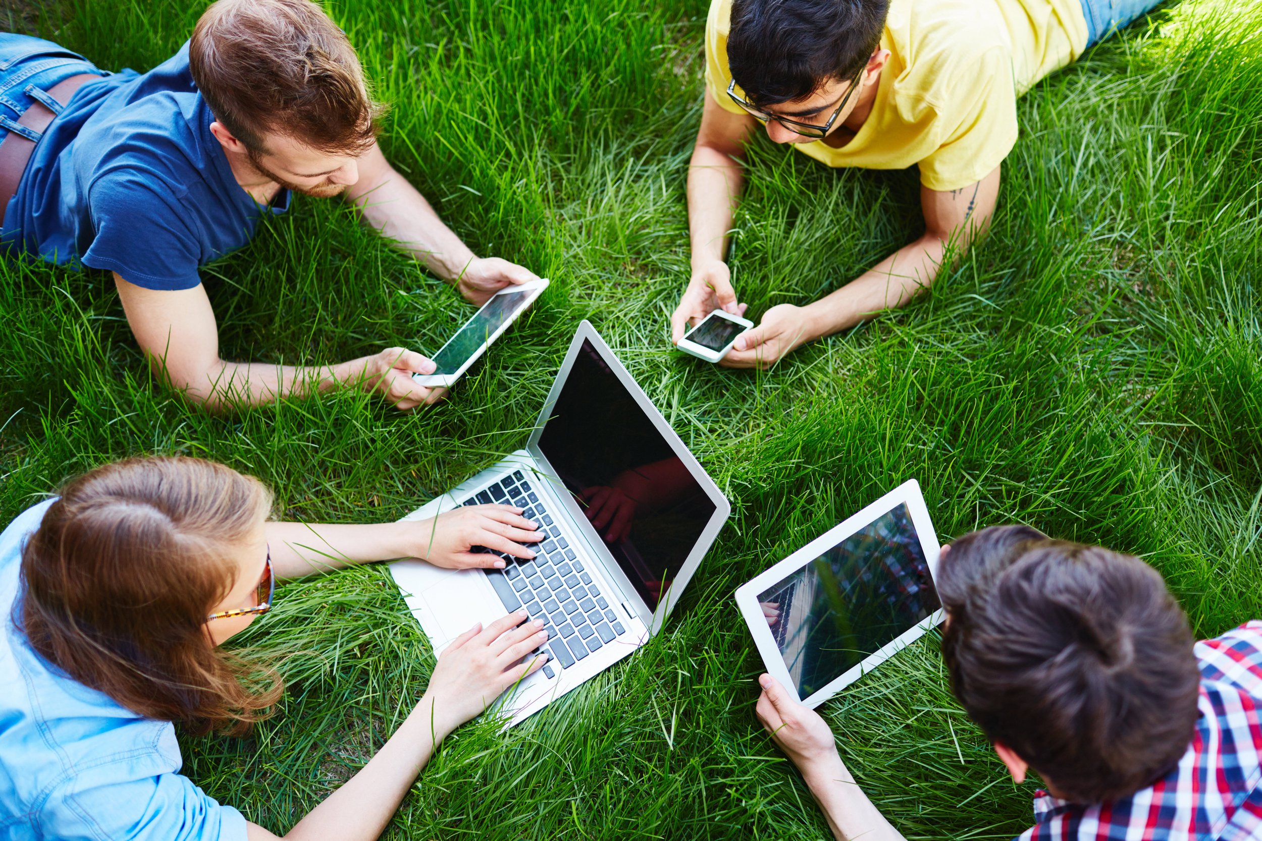 Group of five young people lying on green grass, using laptops, tablets, and smartphones.