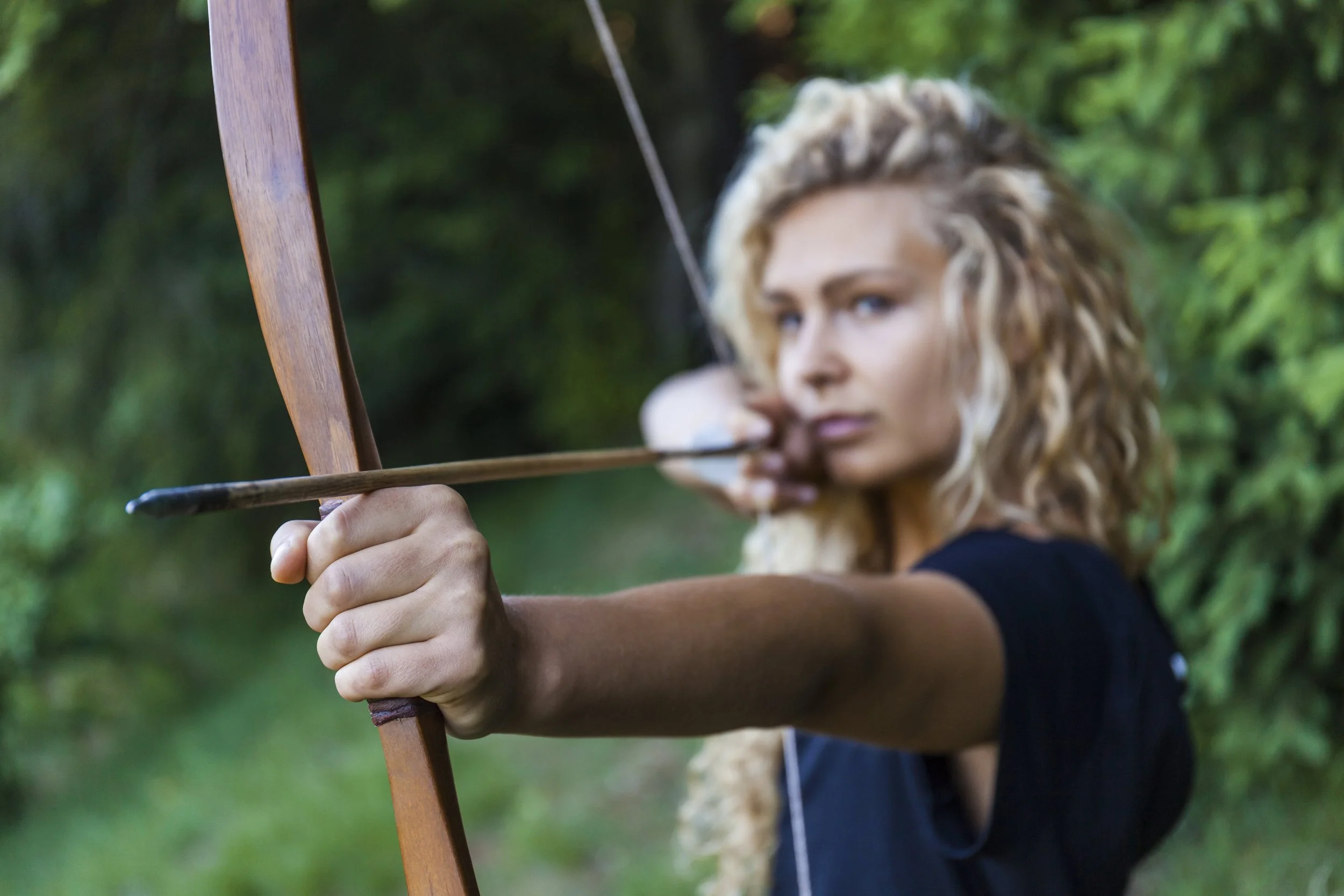 Woman with curly blonde hair drawing a bow in an outdoor wooded area.