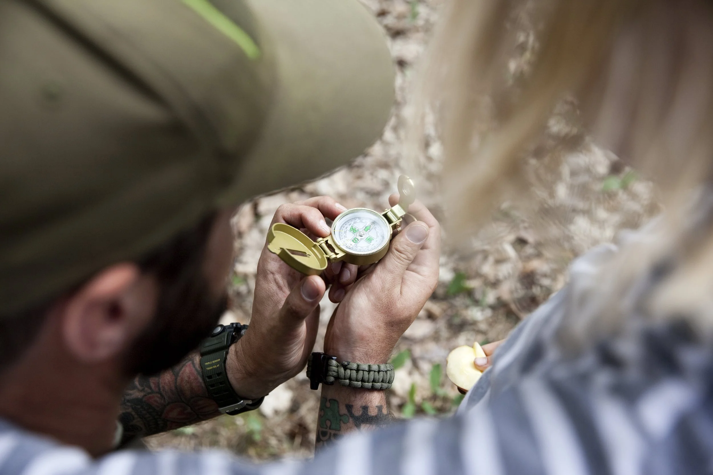 Two people outdoors, one holding a compass and the other holding a mushroom, surrounded by trees and leaves.