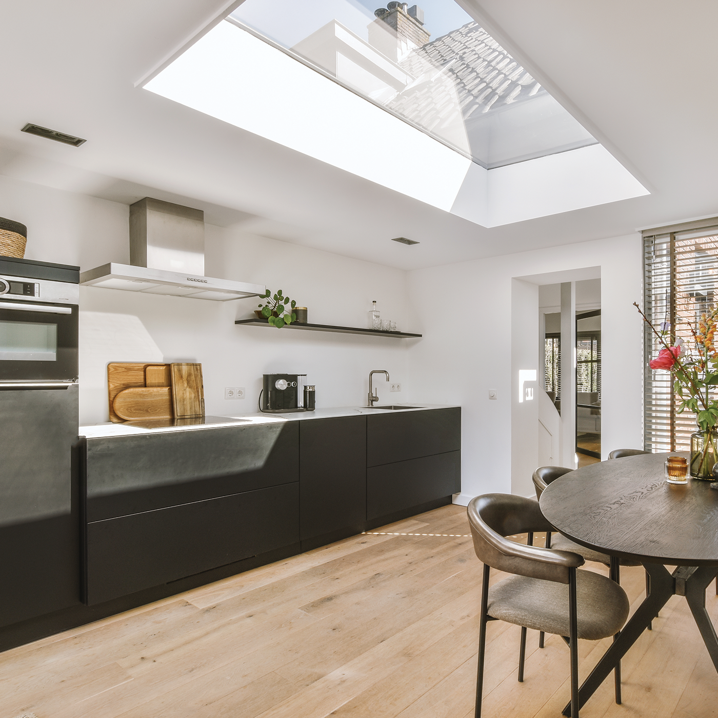 Modern kitchen with black cabinets, wooden floor, and a large skylight letting in natural light. There is a round dining table with chairs and a window with wooden blinds.