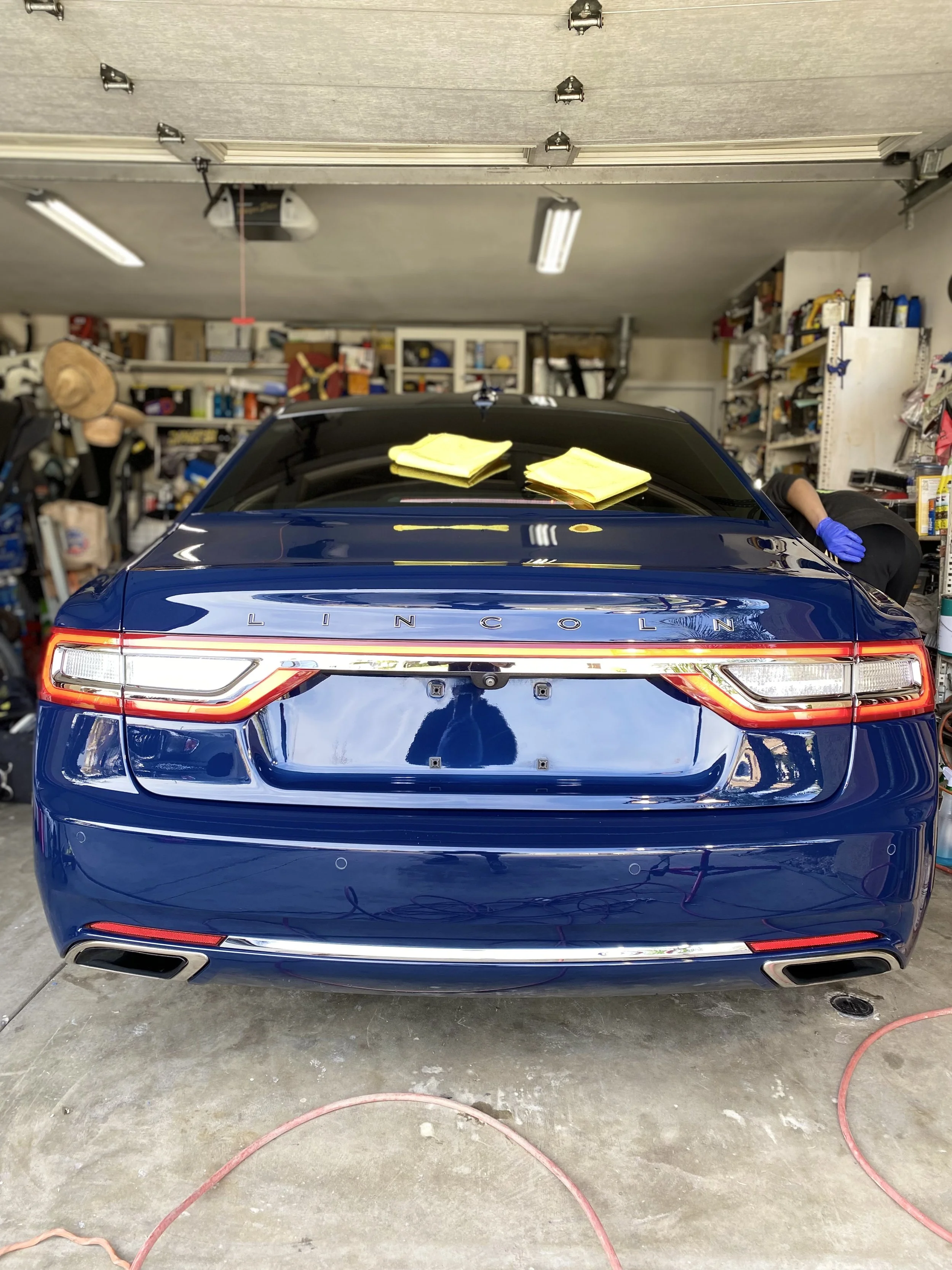 Rear view of a blue Lincoln car in a garage, with yellow cleaning cloths on its trunk and a person in gloves cleaning or working on it.