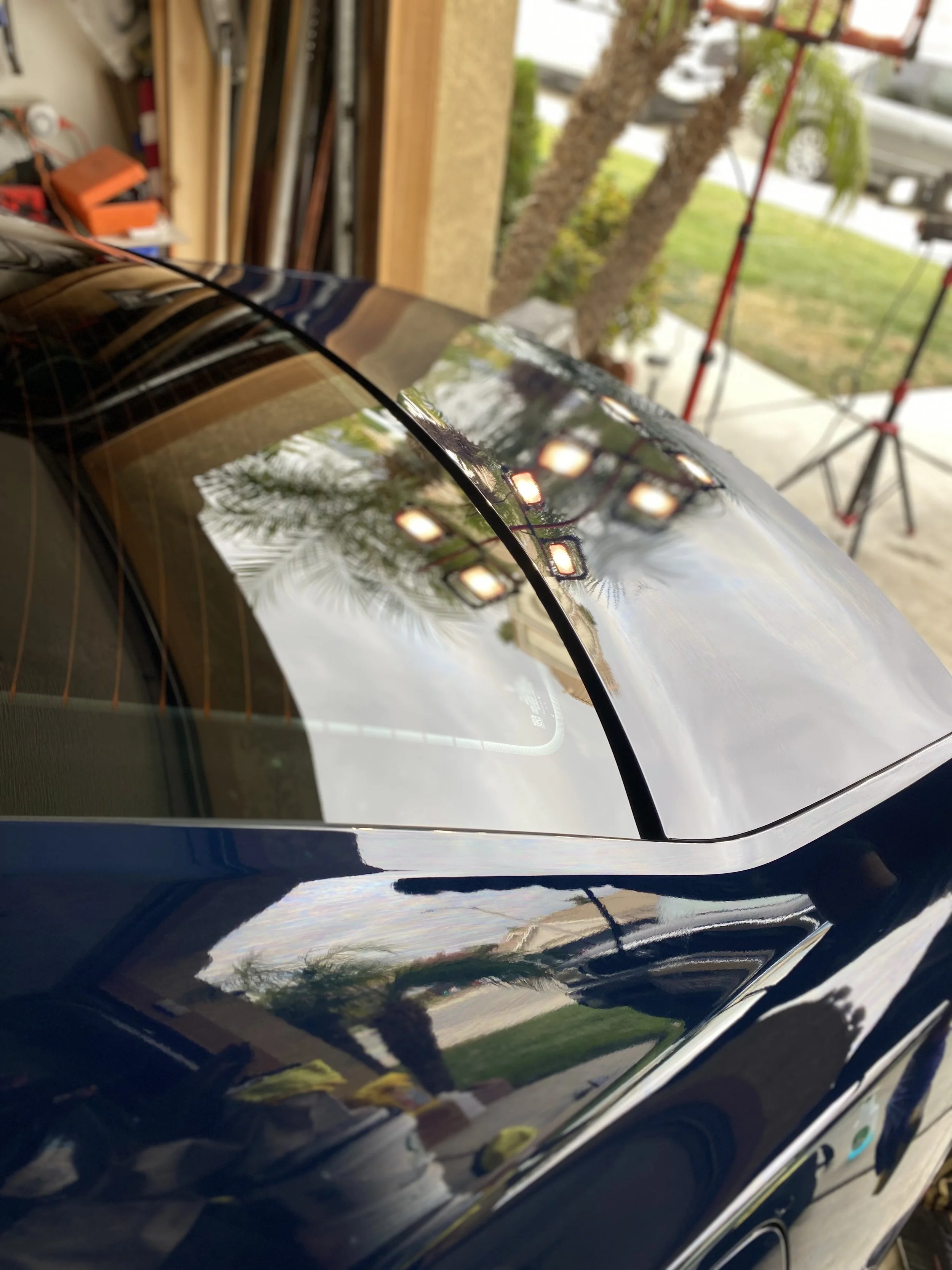 Close-up of a black car's rear window and roof, reflecting trees, clouds, and garage lights, with a garage and outdoor scene in the background.
