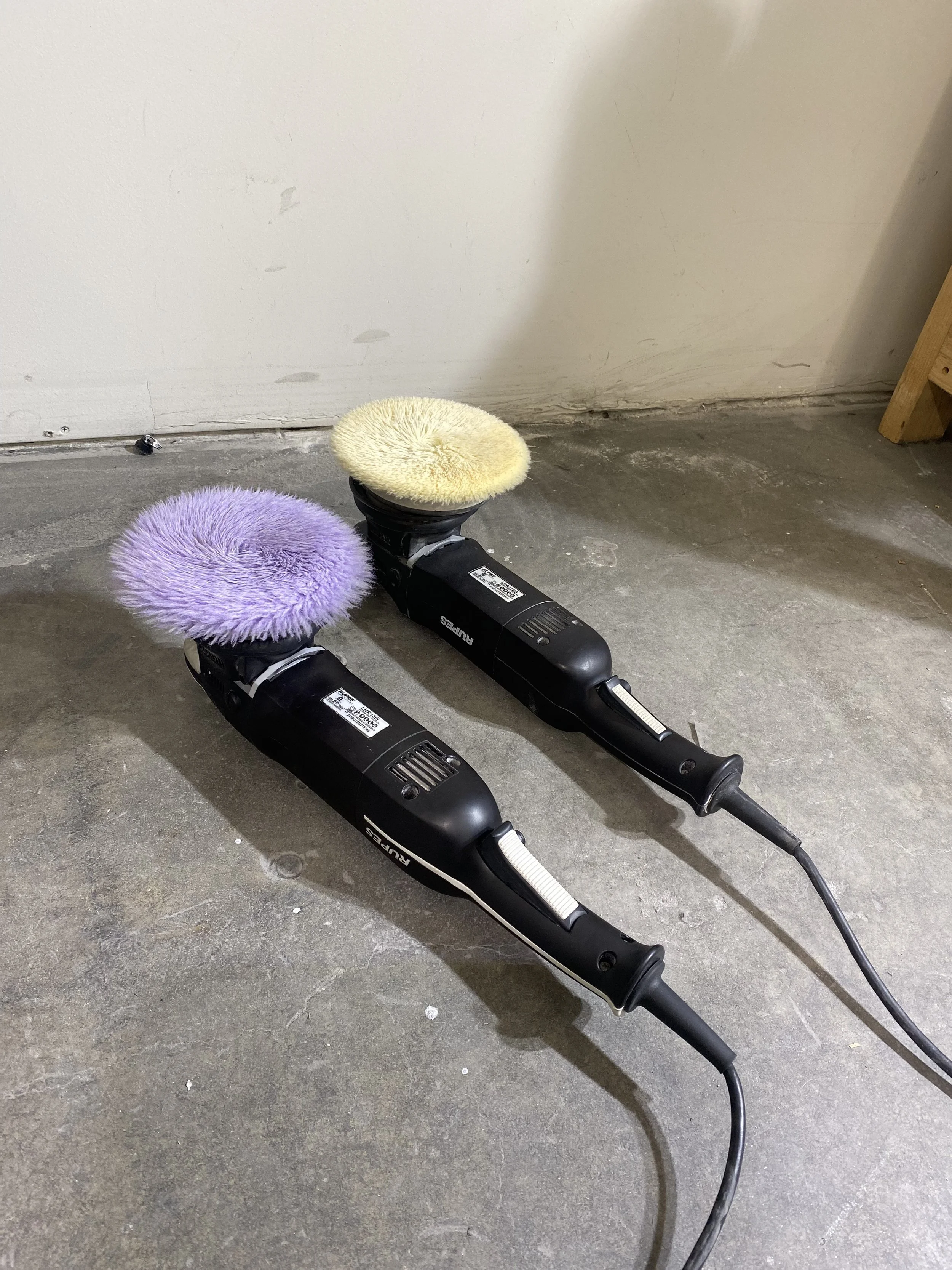 Two electric polisher buffers with fuzzy scrub pads, one yellow and one purple, on a concrete floor next to a white wall.