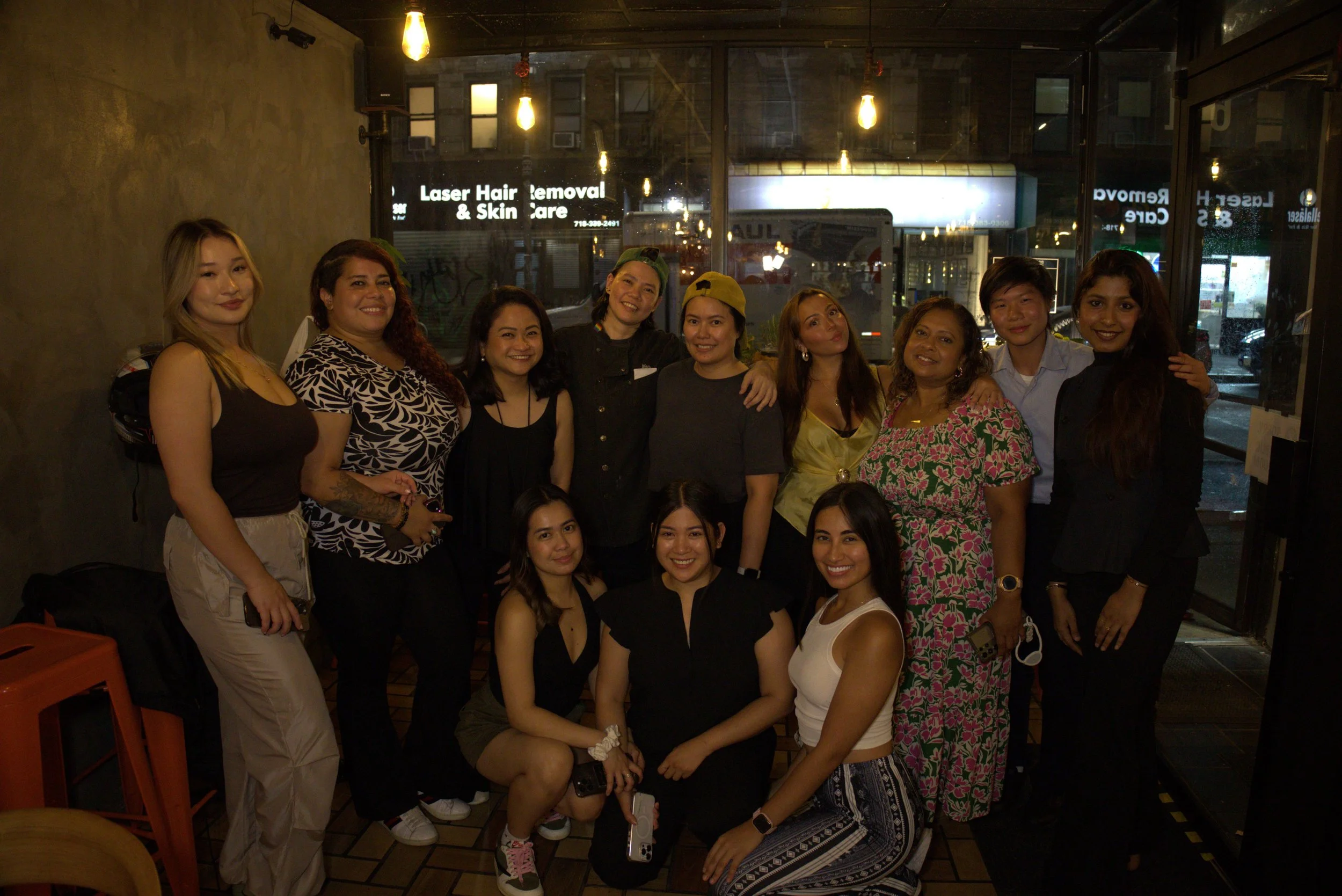 A diverse group of fifteen women gathered in a dimly lit indoor space, posing for a group photo in front of large glass windows with signs for a laser hair removal and skincare clinic in the background.