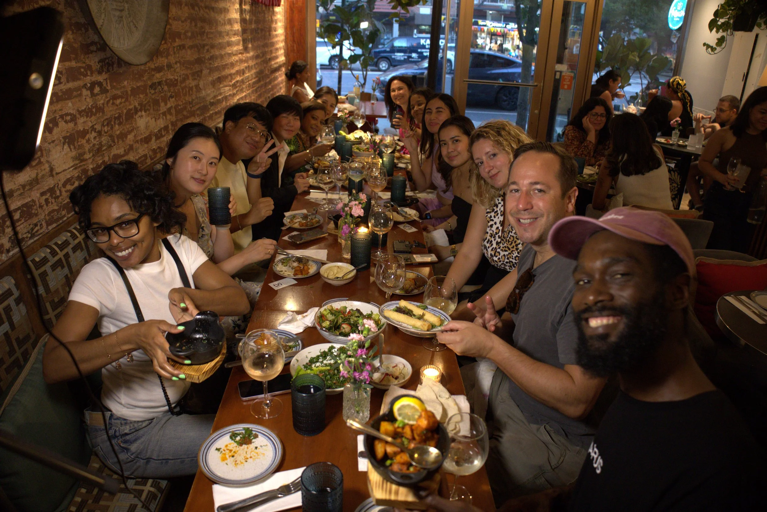 A group of people enjoying dinner at a restaurant, sitting around a long table with plates of food, drinks, and flowers in a cozy, well-lit setting.