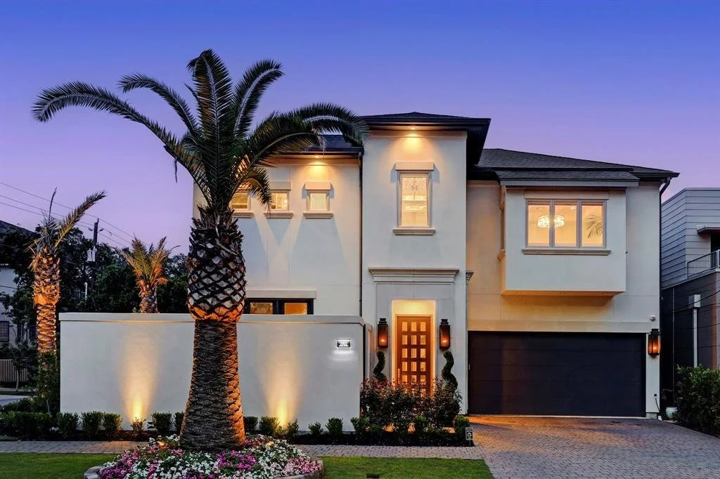 Modern two-story house with beige walls, large front window, a decorative front door, and a two-car garage, surrounded by palm trees and landscaping during twilight.