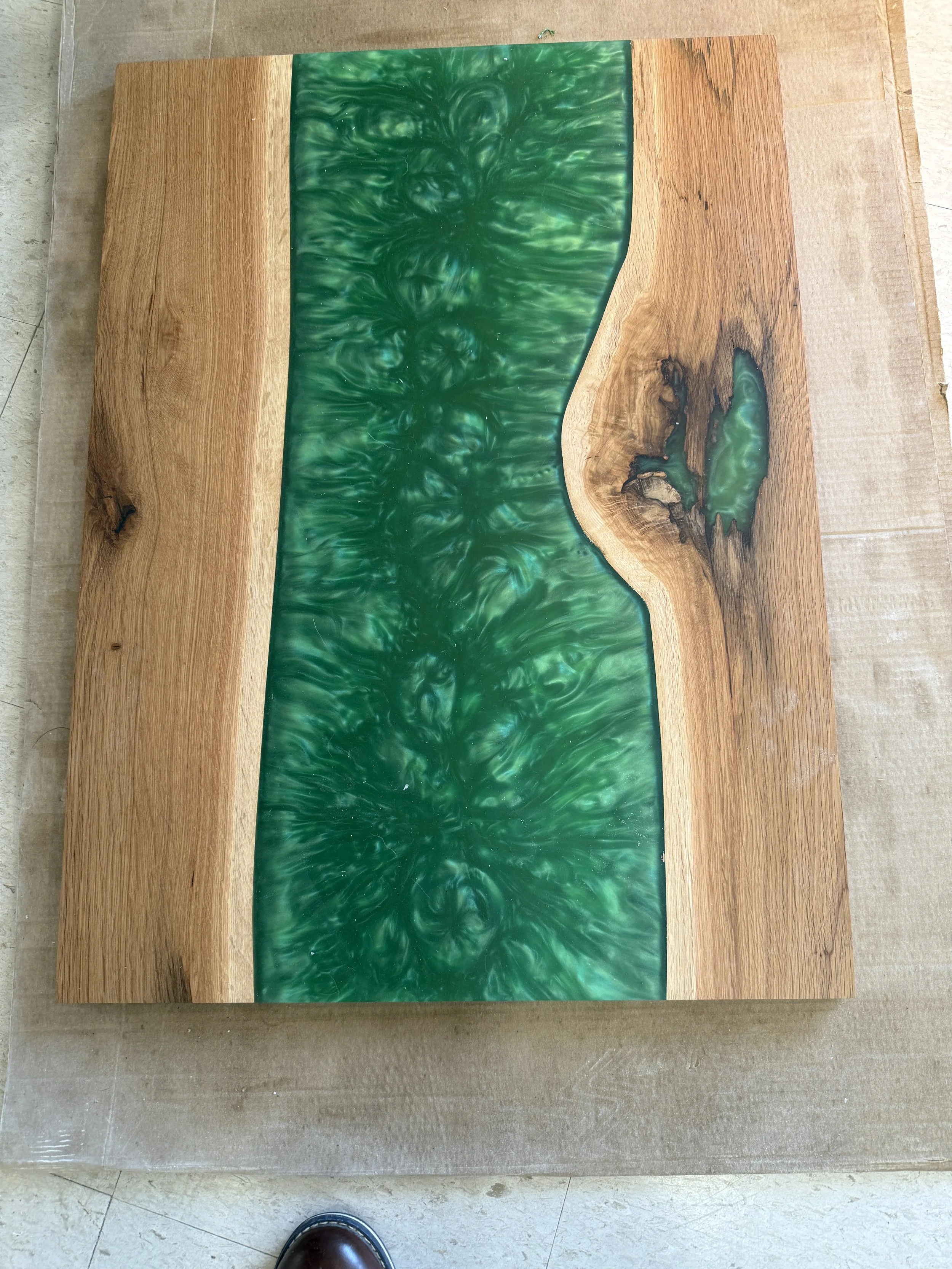 A wooden table with a green resin inlay design. The inlay has a swirling pattern and is bordered by natural wood on three sides, with a more irregular edge on one side.