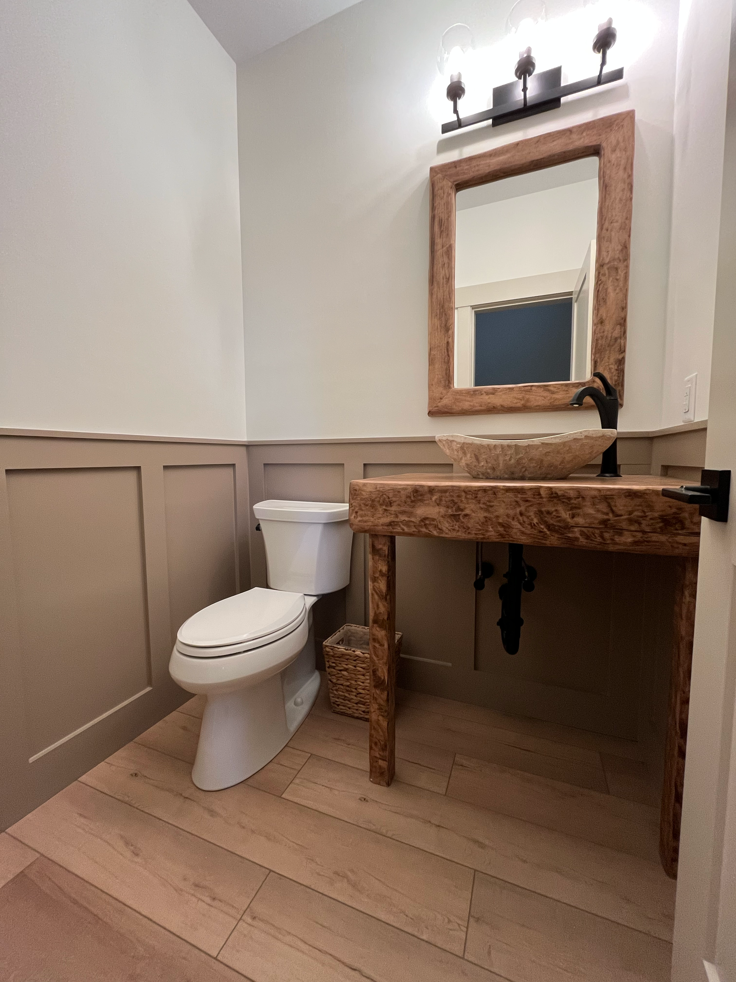 A rustic bathroom featuring a wooden vanity with a stone vessel sink, a black faucet, a large mirror with a wooden frame, a modern light fixture above the mirror, white walls, a white toilet, and a woven wastebasket on the wooden floor.