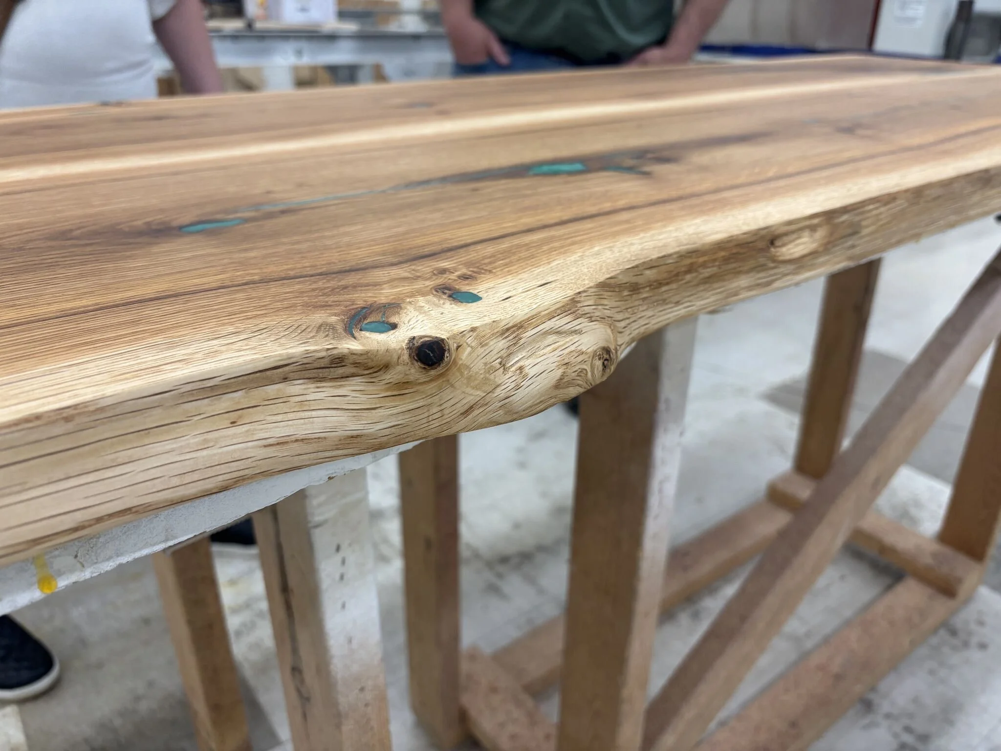 Close-up of a wooden table with visible grain, knots, and a slight natural edge, supported by a wooden frame amid a workshop setting.