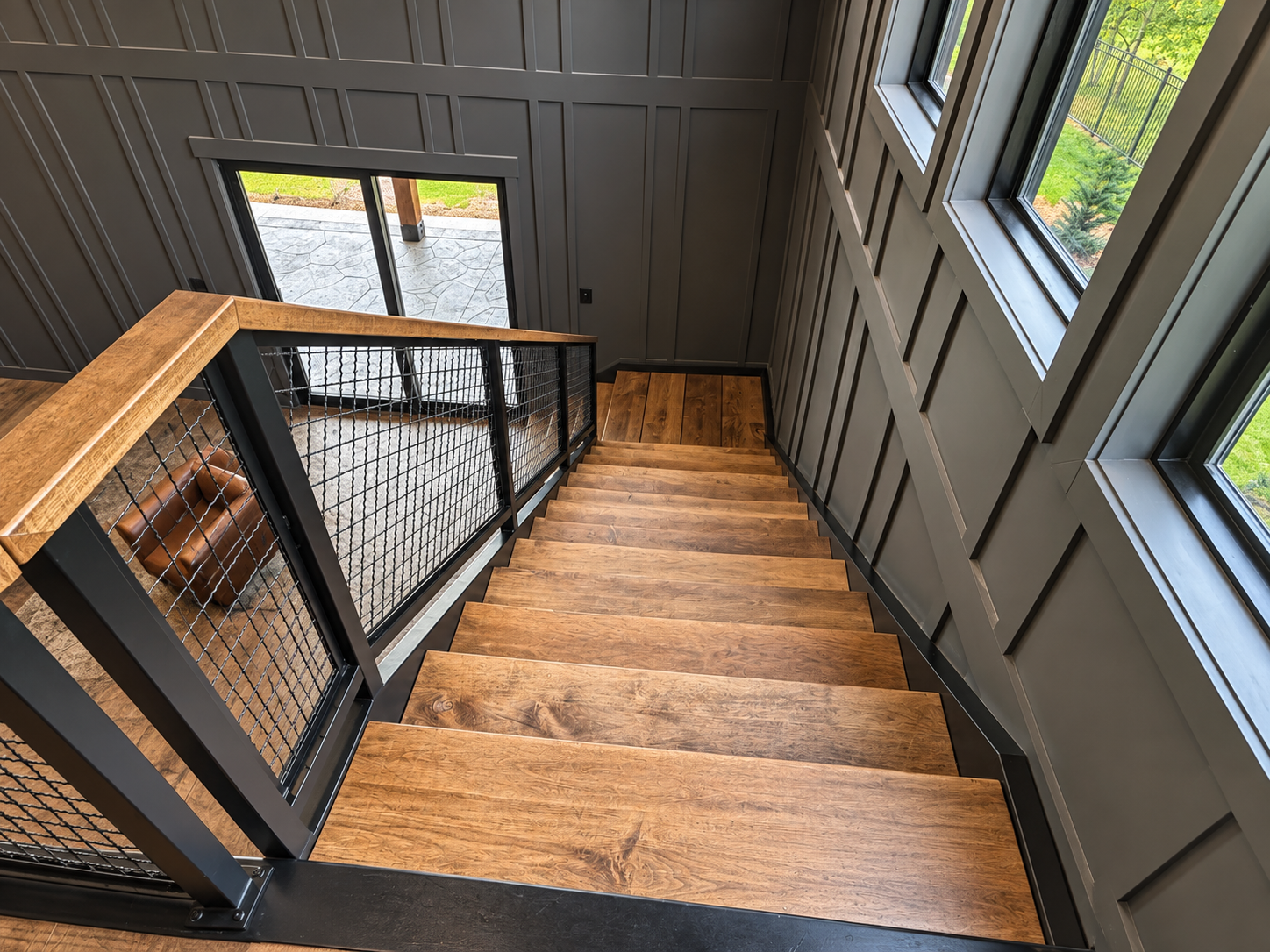 Wooden staircase inside a house, with large windows on the right and a gray wall on the left, leading down to a small landing with sliding glass door, overlooking a backyard with a stone patio and green grass.