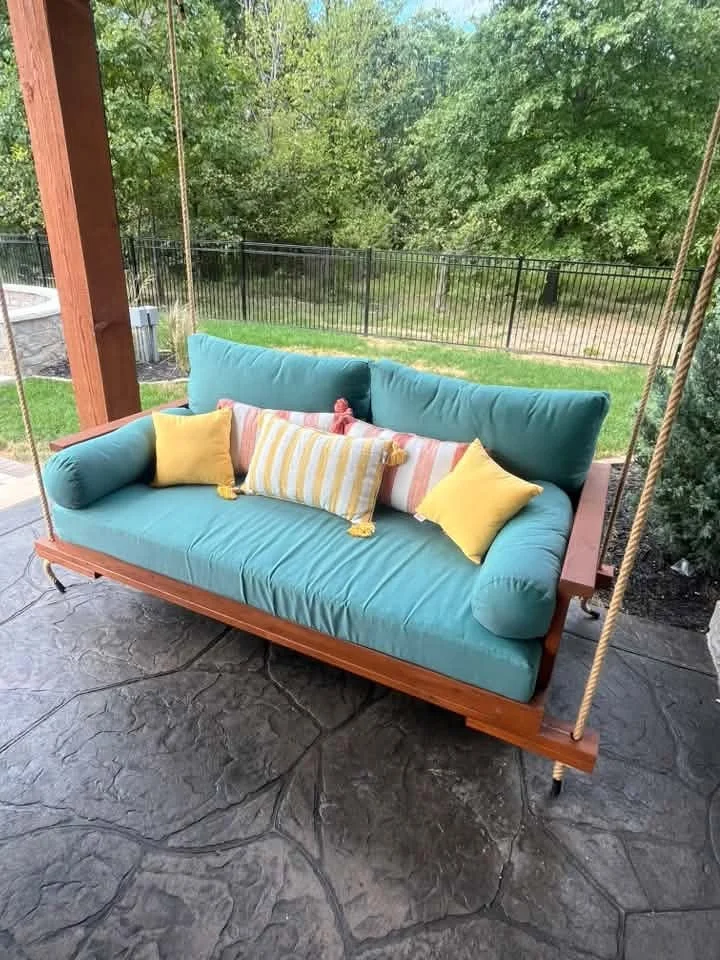 Outdoor wooden swing with teal cushions and decorative pillows on a screened porch, overlooking a green backyard with trees and a black fence.
