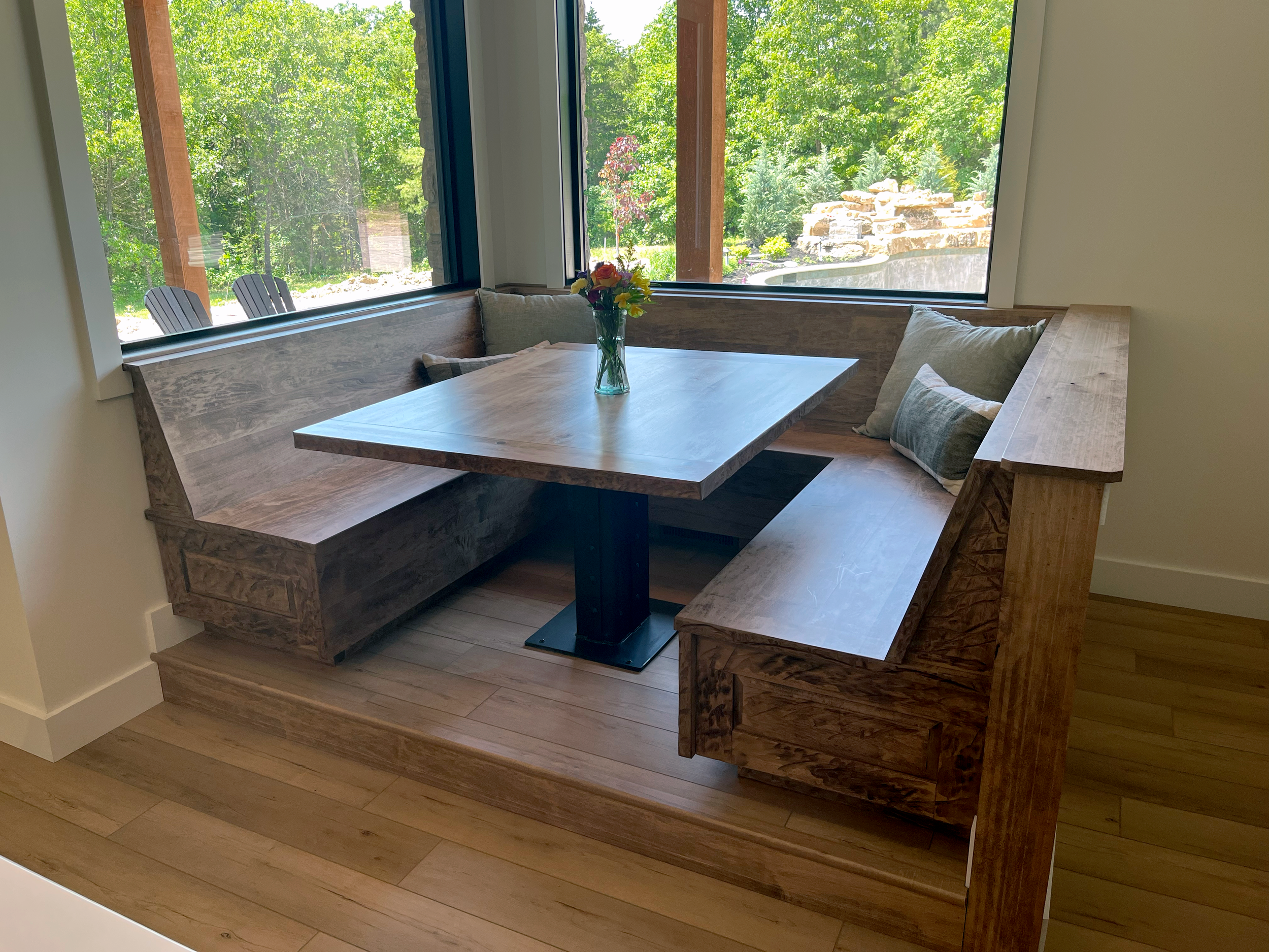 Wooden corner bench with cushion and flower vase near large windows showing greenery outside.