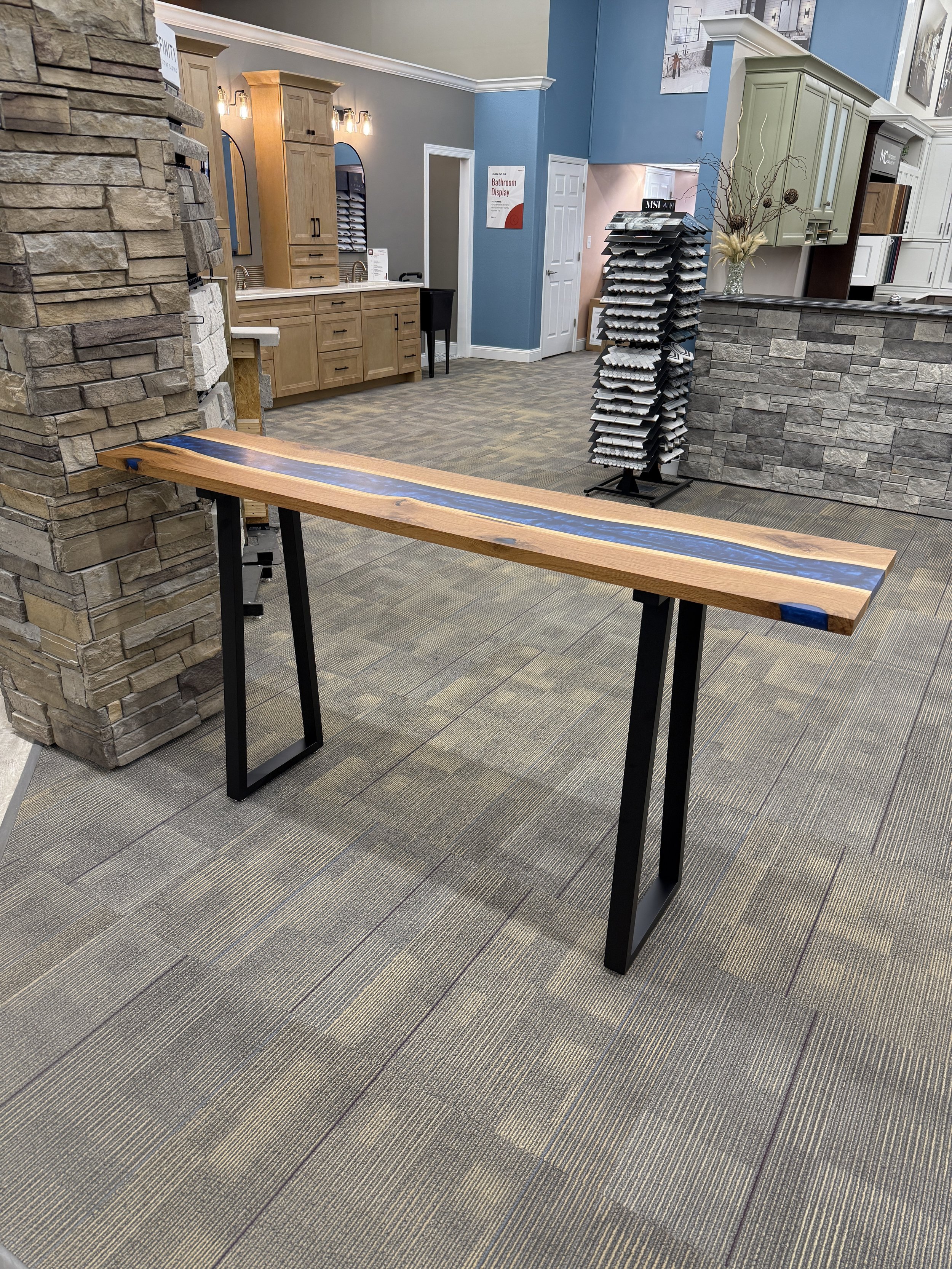 A wooden table with a blue resin river running through the center, supported by black metal legs, in a showroom with stone and wood decor.
