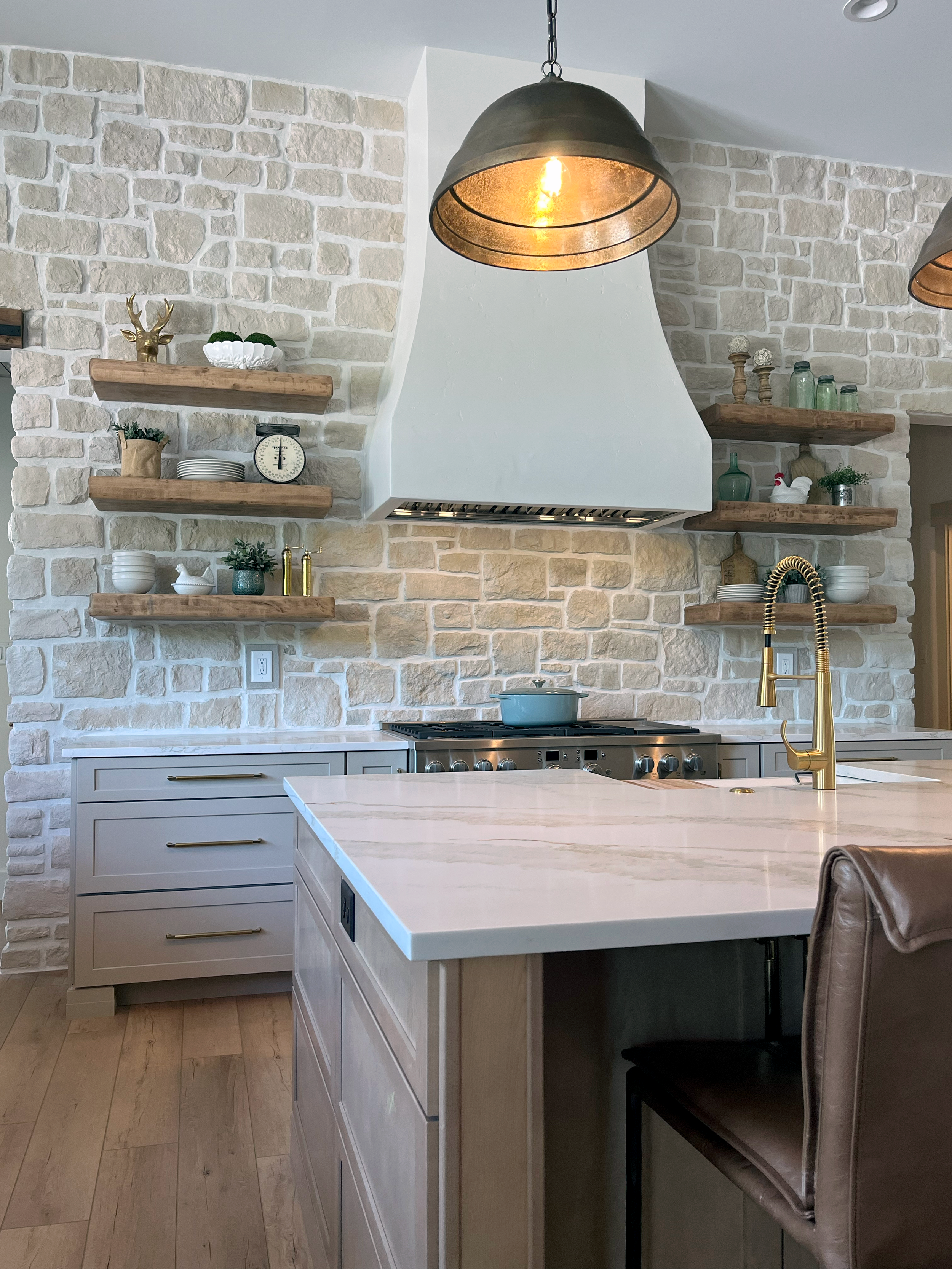 A modern kitchen with a white island, gold faucet, open wooden shelves on a textured brick wall, and a white range hood with warm pendant lighting.