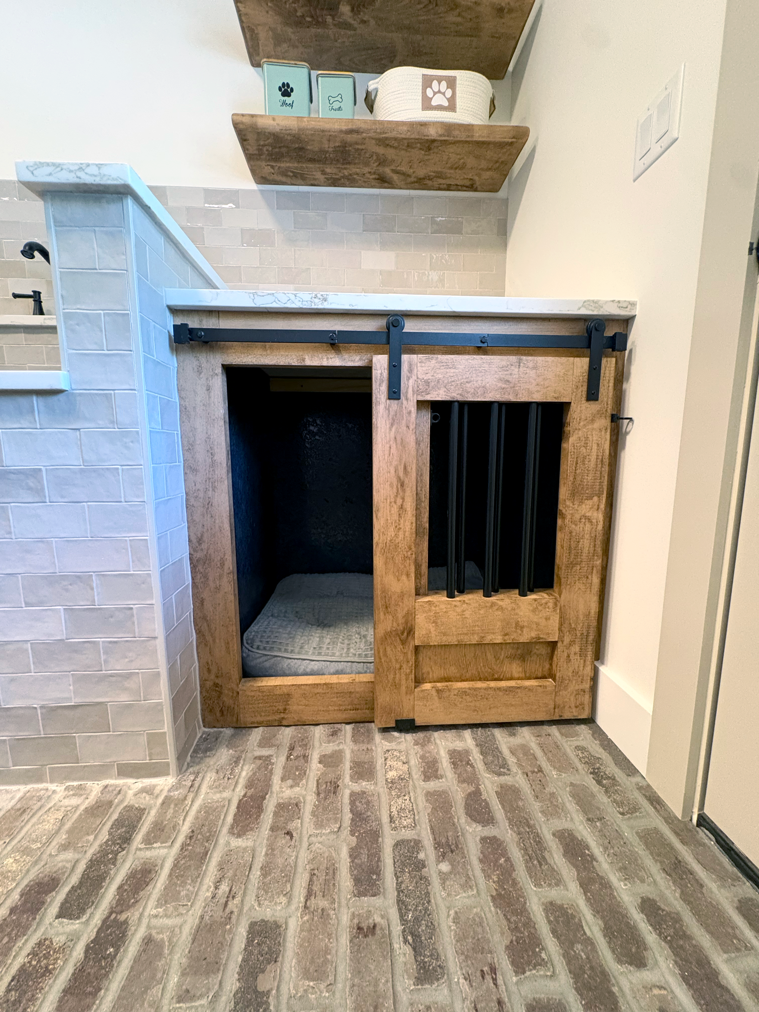 Interior of a laundry or utility room with a brick floor, a wooden cabinet with a black sliding barn door, a shelf with pet-themed storage containers, and a tiled wall with a small sink.