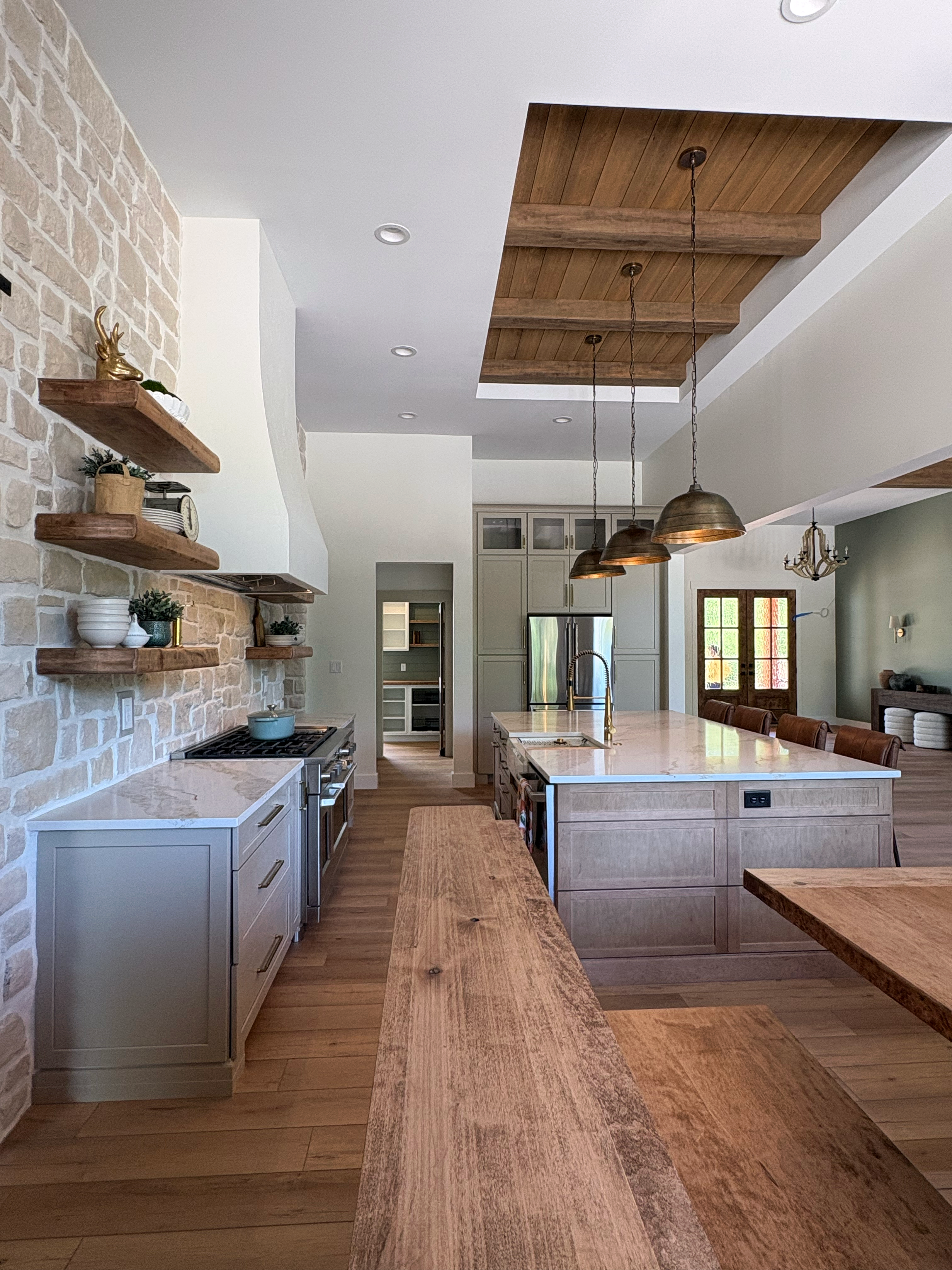 Open kitchen with wooden beams, pendant lights over a marble island, floating shelves on a feature wall, and a view into a dining area with a chandelier.