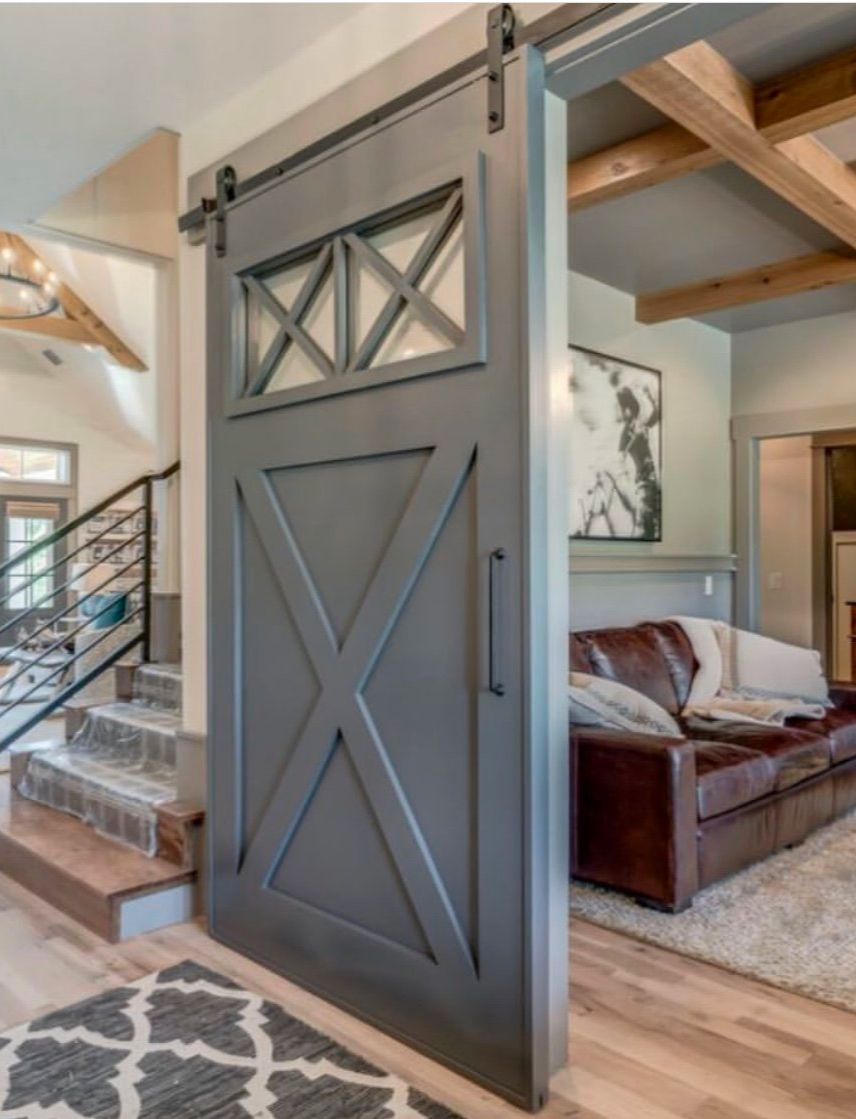 A gray sliding barn door with buckle hardware, partially open, revealing a living room with a brown leather sofa and abstract wall art.