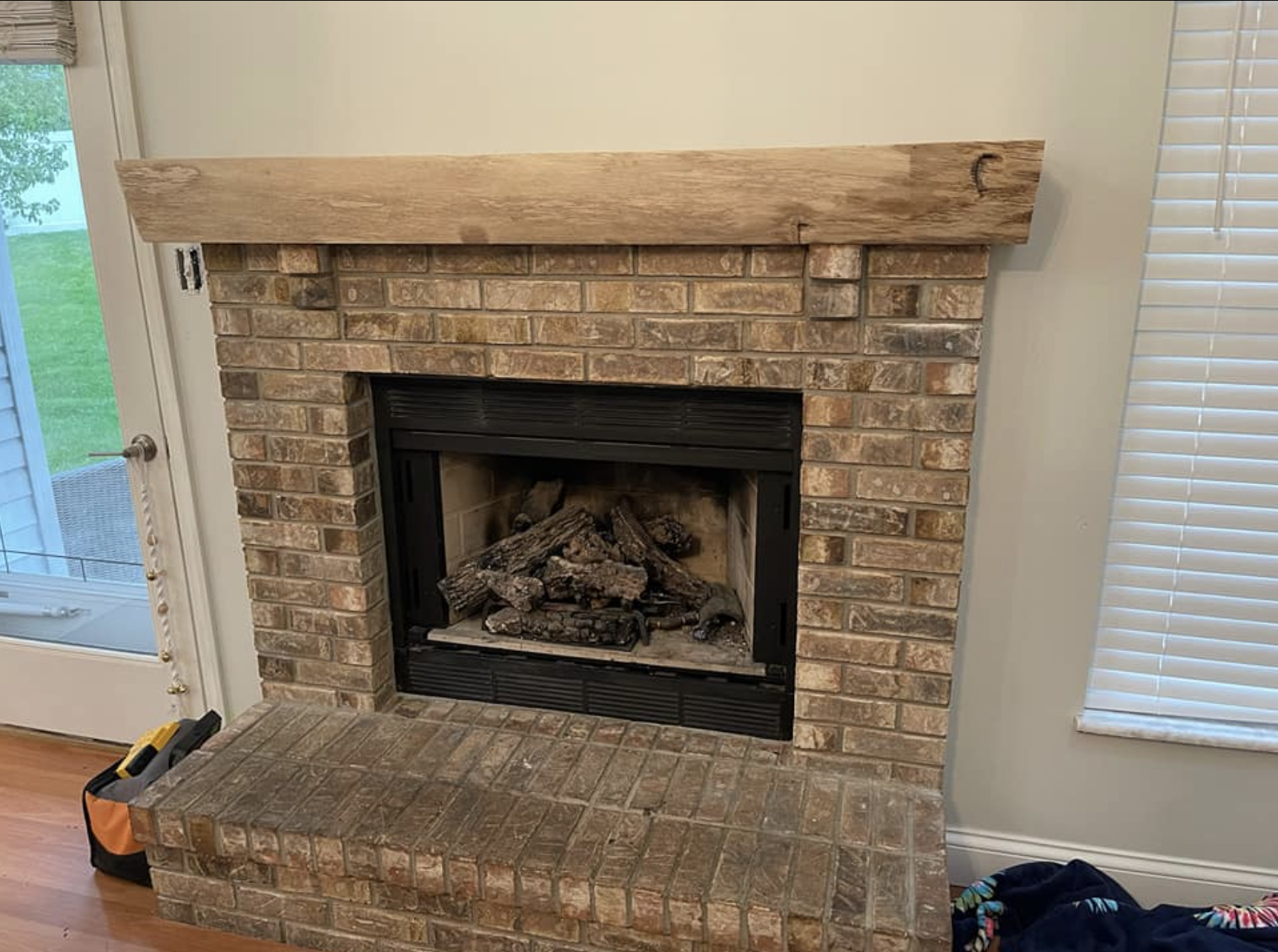 Brick fireplace with a wooden mantel and gas log insert in a living room, with a sliding glass door to the left and a window with blinds to the right.