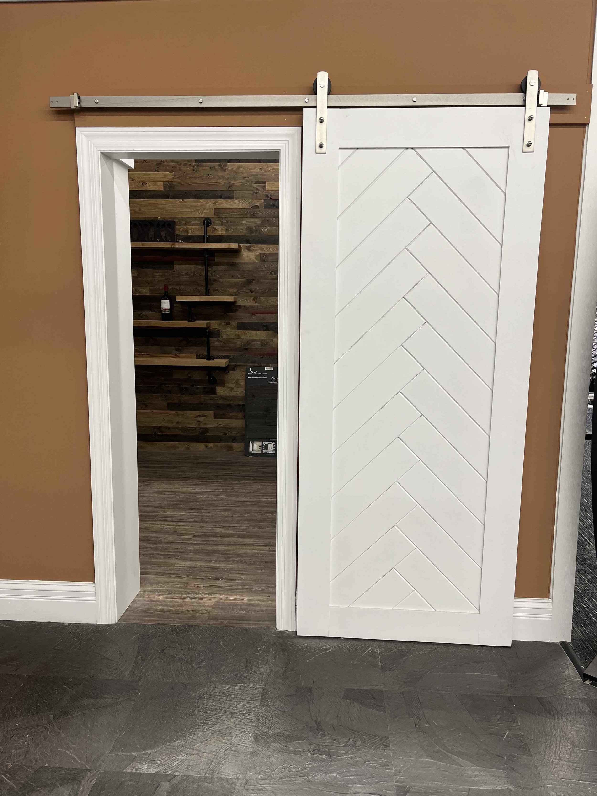 A sliding barn door with a white chevron pattern, on a brown wall, partially open to reveal a room with dark wood plank walls and shelving. The sliding door is mounted on a metal track.
