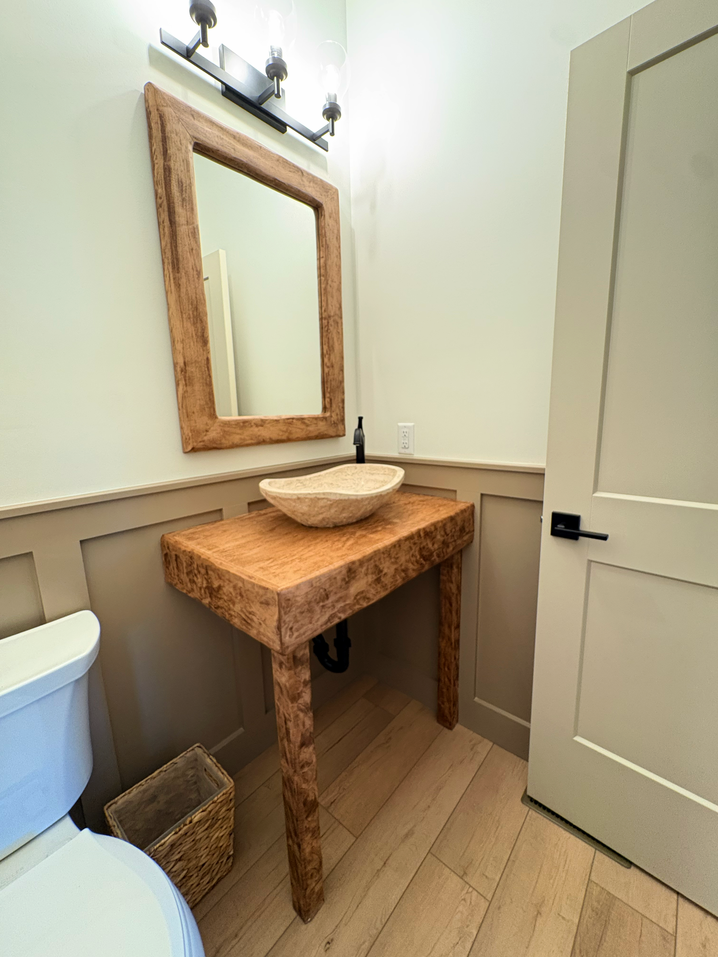 Interior of a small bathroom with a wooden vanity holding a stone vessel sink, a large mirror with a wooden frame, a white toilet, a woven basket underneath the vanity, and a gray cabinet on the right side of the image.