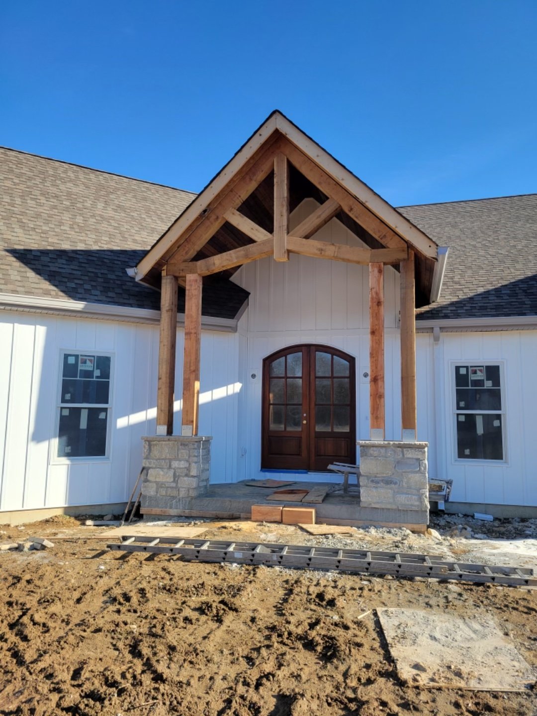 Under construction house with a front door, white siding, and a gable roof, featuring a wooden porch frame and stone foundation steps, on a dirt lot under a clear blue sky.