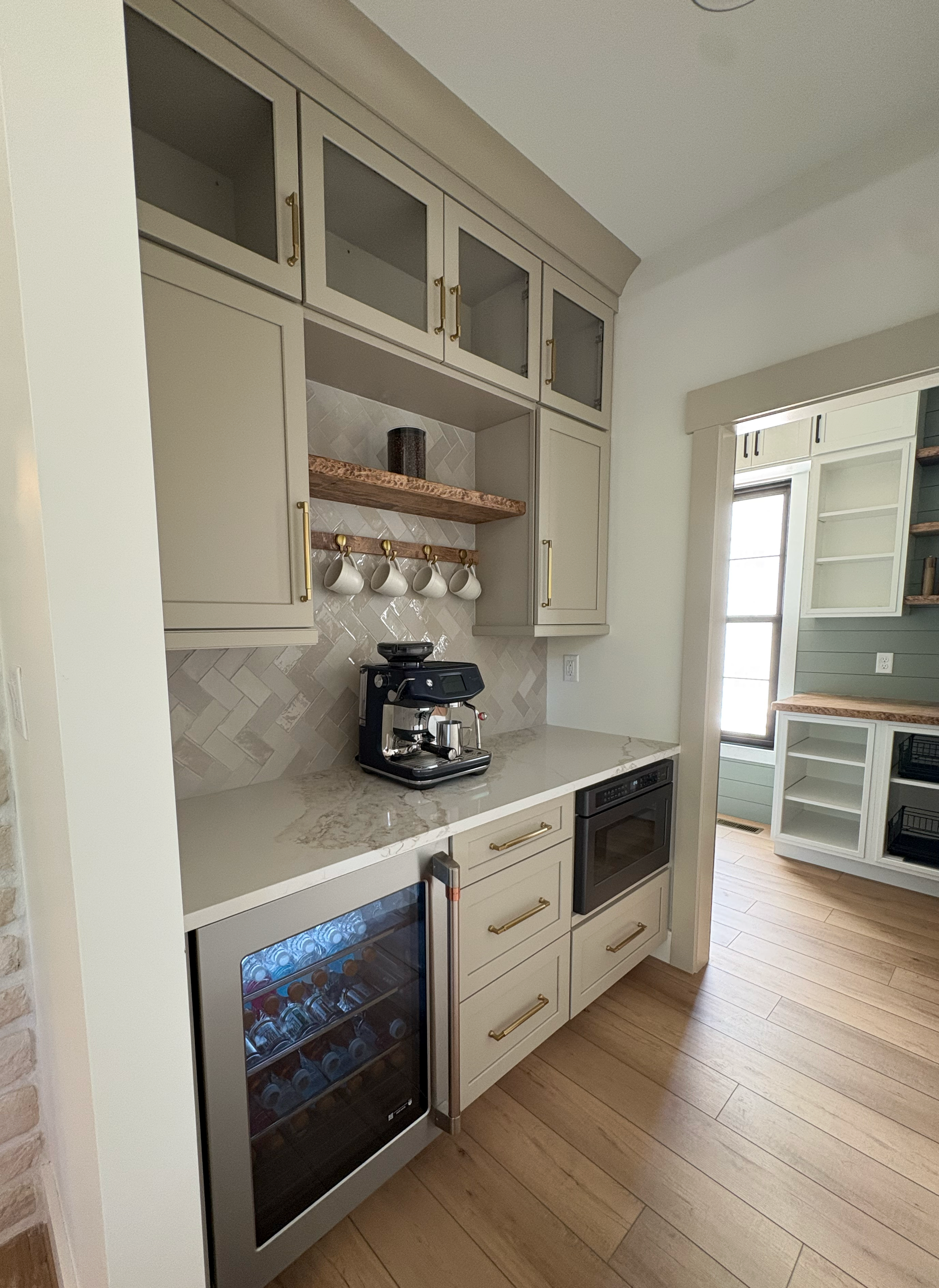 A modern kitchen with beige cabinets, a marble countertop, a wooden shelf with white mugs, a coffee machine, and a wine cooler, with a window in the background.