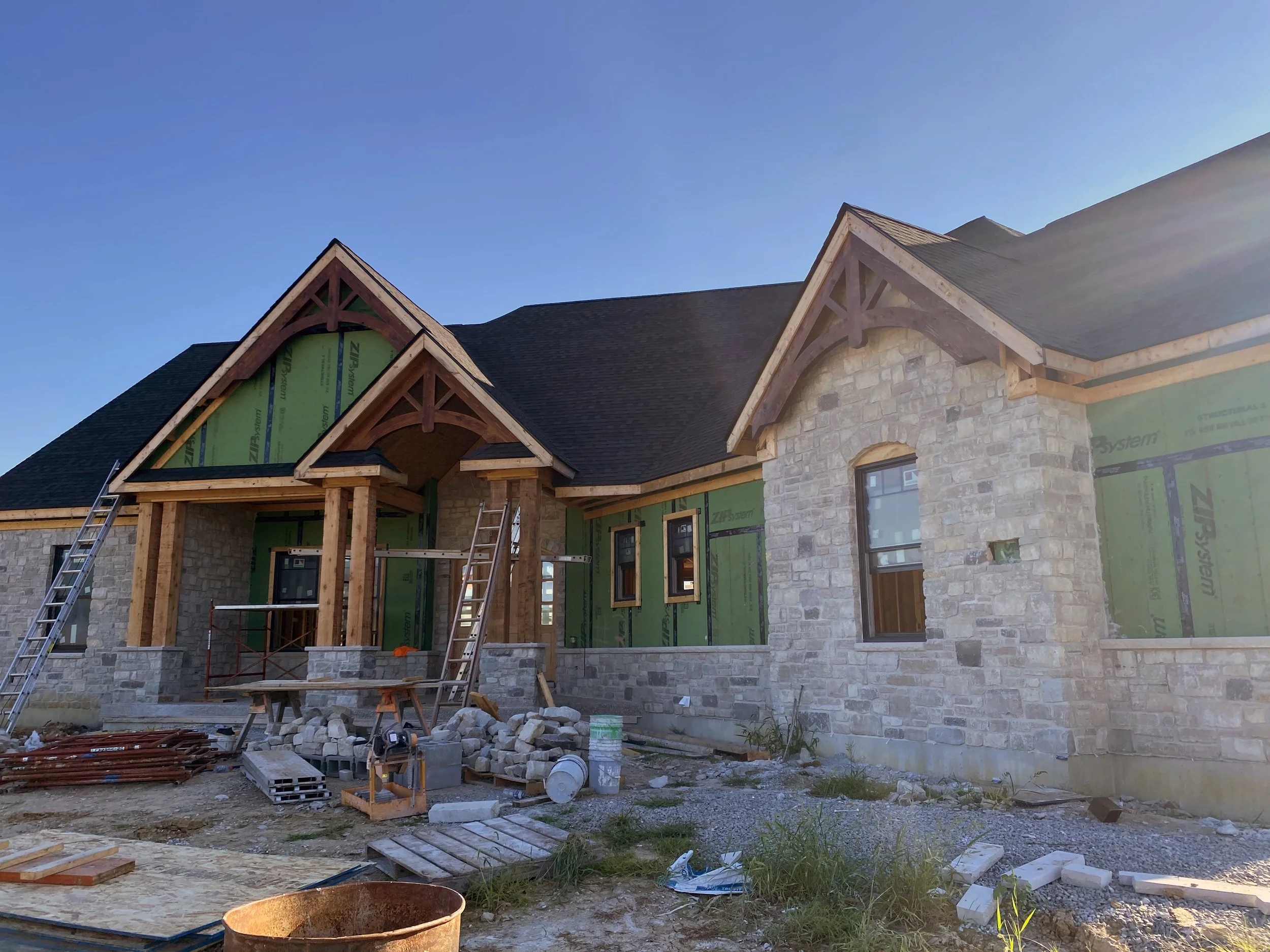 A house under construction with a stone facade, wooden framing, and green insulation panels, featuring multiple windows and a steep roof with gables.