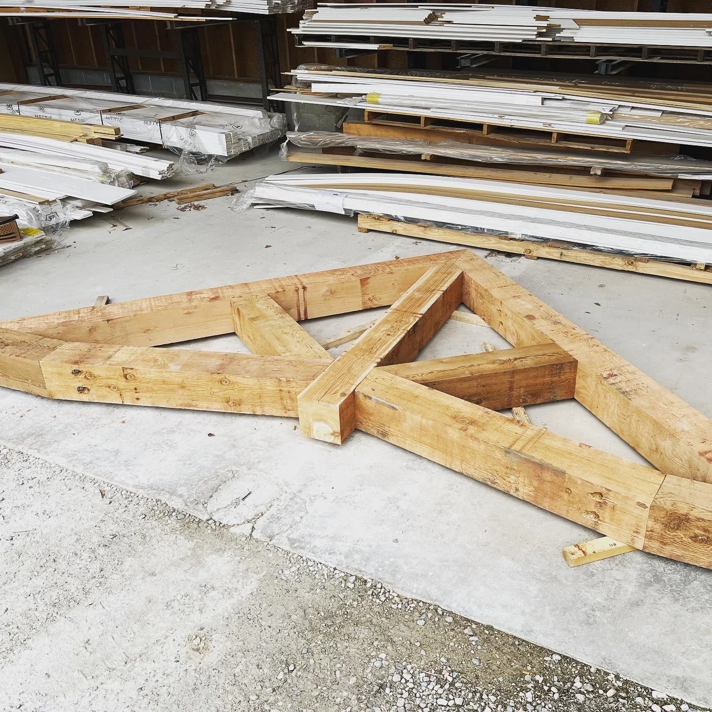 A large wooden sign frame lying on the concrete floor at a construction site with stacked wood panels and trims in the background.