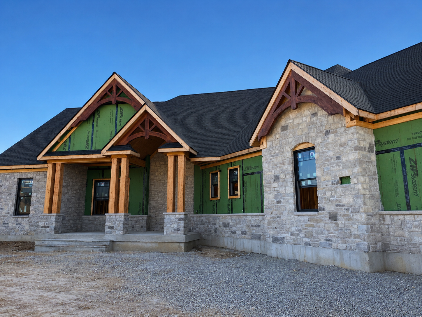A house under construction with stone walls, wooden beams, and a black shingled roof, set against a blue sky.
