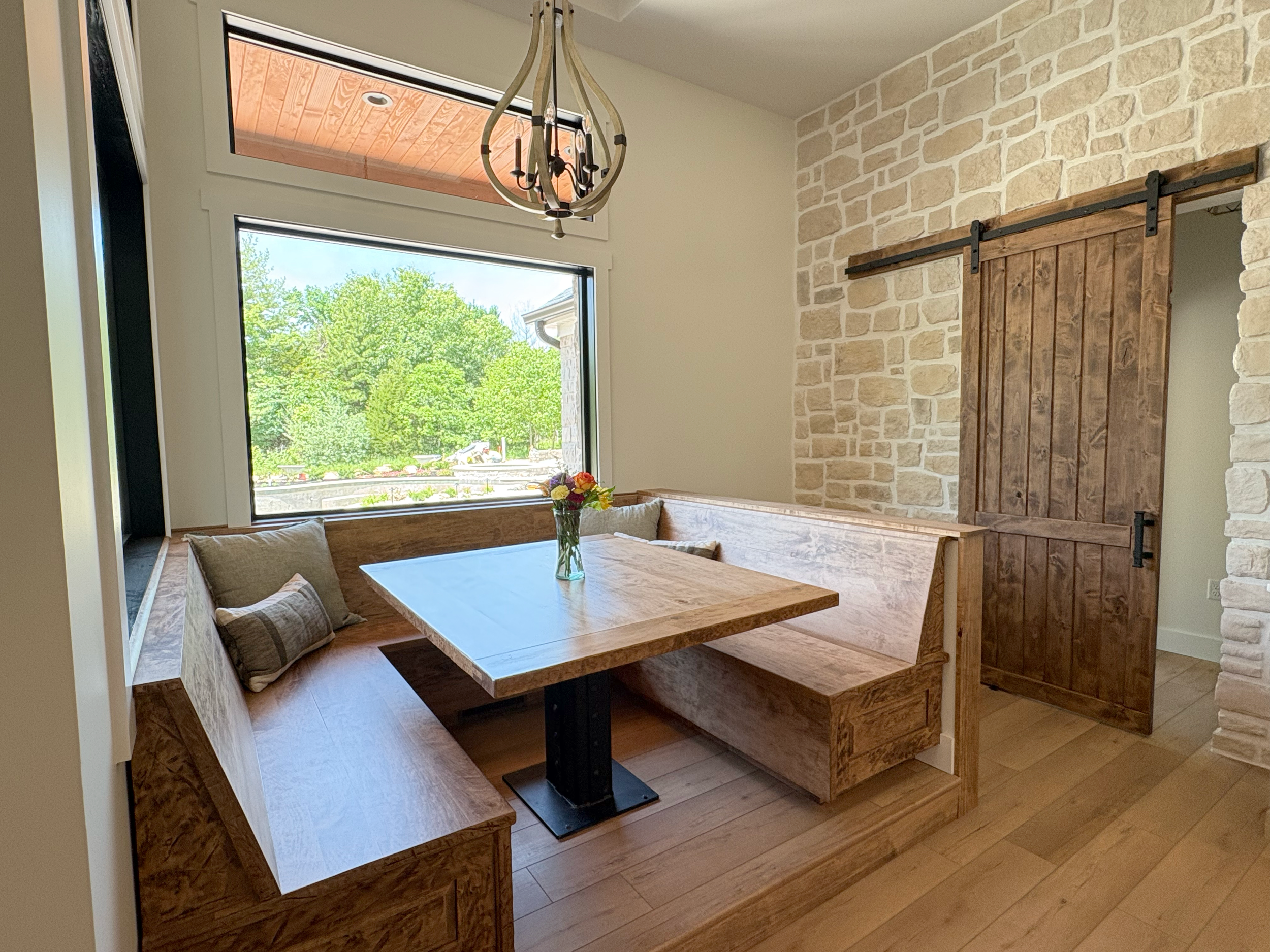 A cozy dining nook with a built-in wooden bench, a wooden table, a window showing green trees outside, a flower vase on the table, and a sliding barn door on a stone wall.