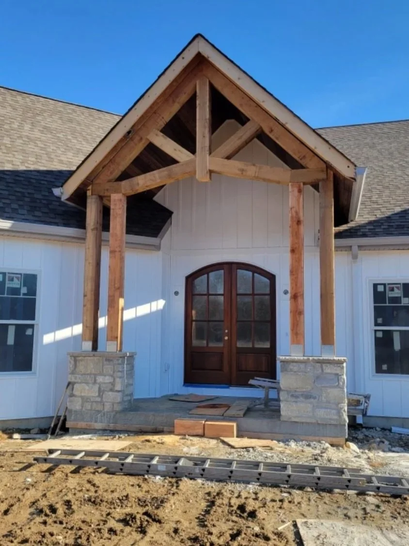 Front entryway of a house under construction with a wooden porch roof frame supported by stone pillars and a wooden front door.