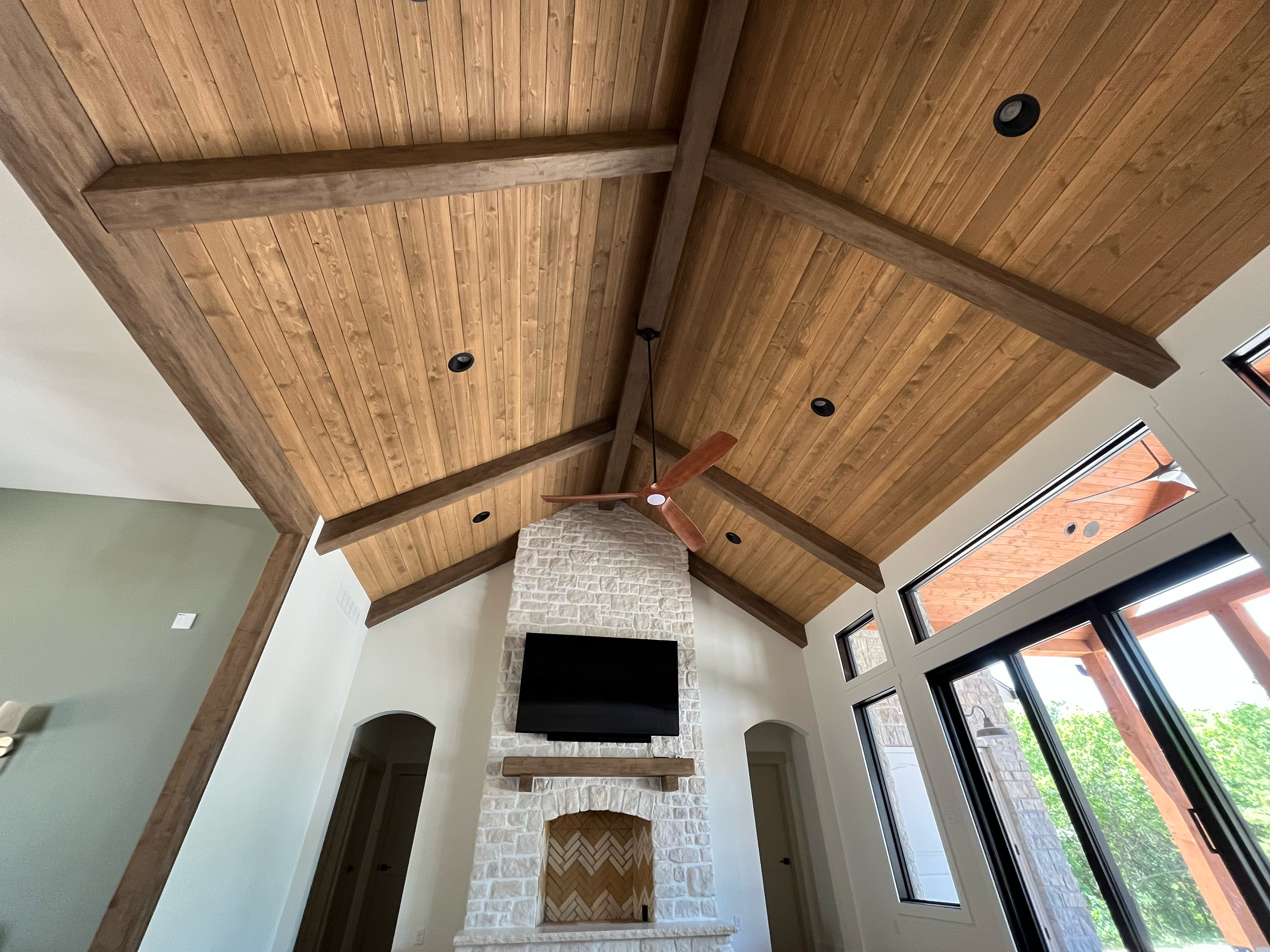 Interior view of a living room with a wooden ceiling, a white stone fireplace, a wall-mounted TV, and large windows with black frames showing greenery outside.