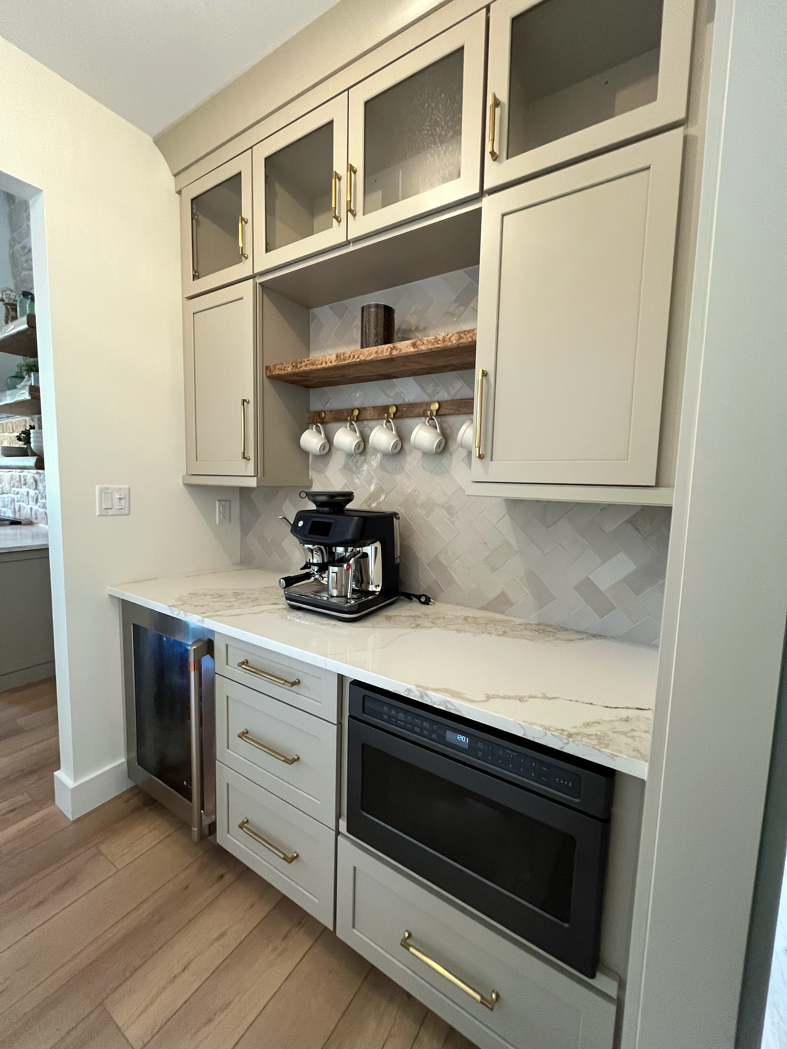 Kitchen area with white cabinets, a marble countertop, a black espresso machine, a small refrigerator, and a built-in oven. There is a wooden shelf with cups hanging underneath and a decorative vase on the shelf.