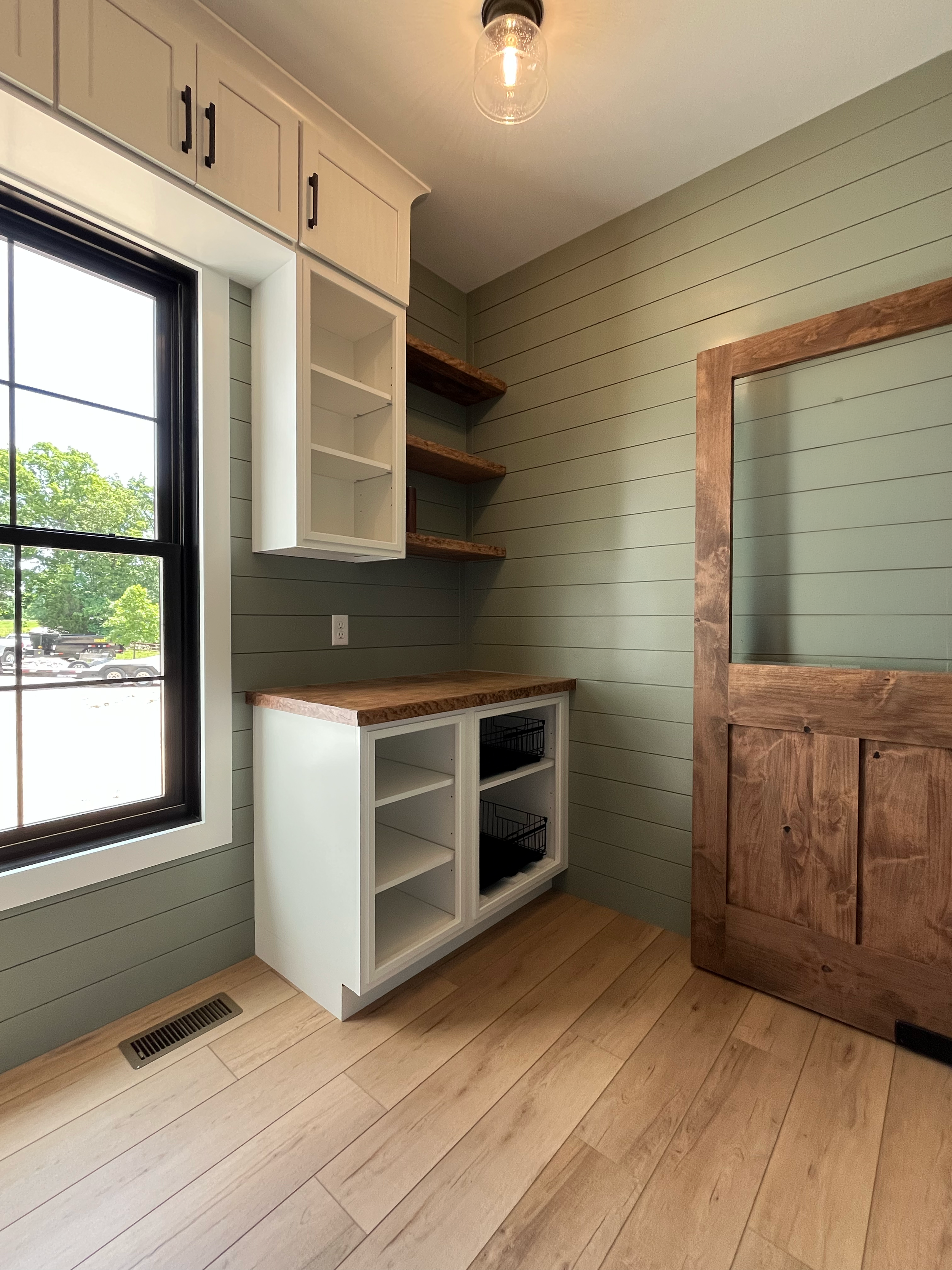 Corner of a room with green shiplap walls, light wood flooring, a window with black trim, and built-in furniture including white cabinets with open shelving and a wooden countertop. There are also brown wooden shelves on the wall and a large wooden-f
