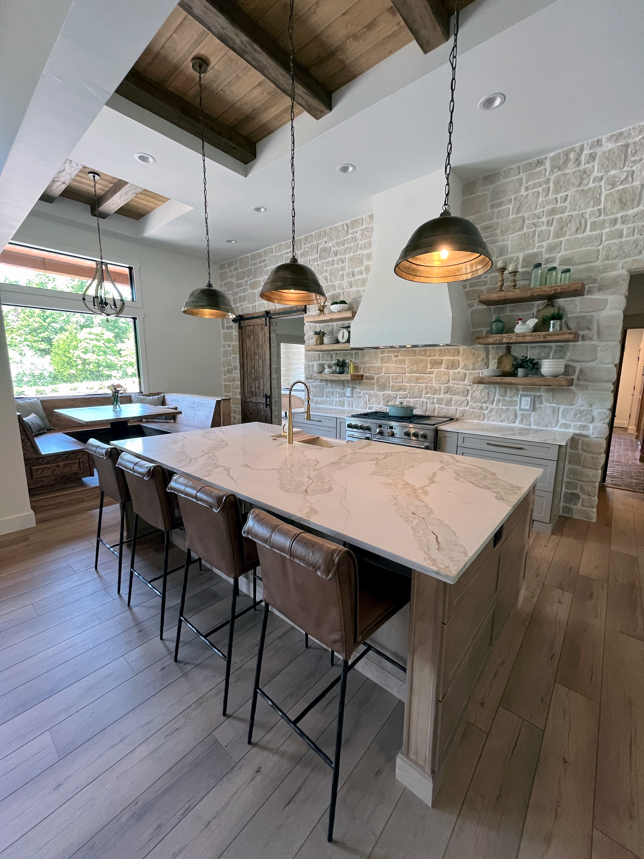 A modern kitchen with a marble island, black barstools, a stone backsplash, floating wooden shelves with jars and decor, black hanging pendant lights, and a window showing green trees outside.