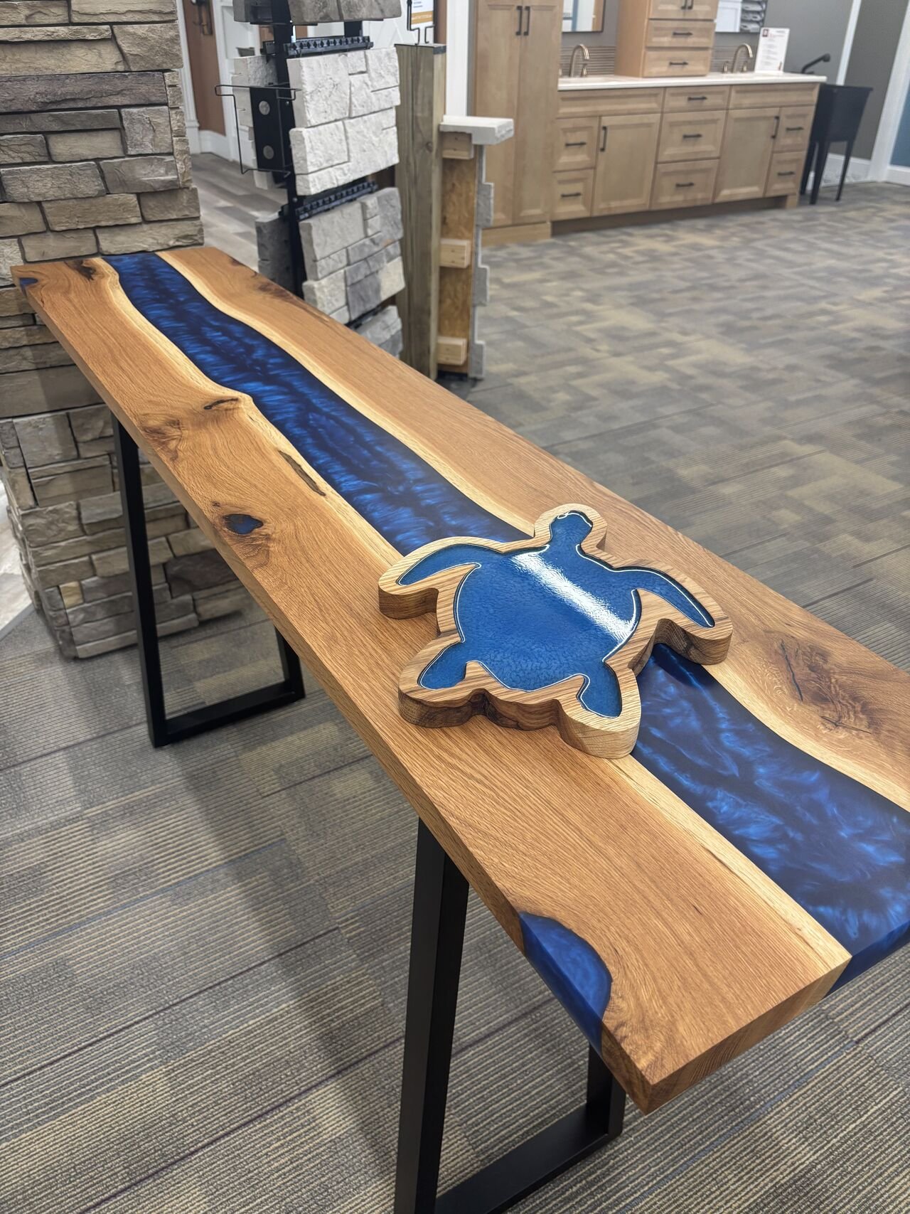 Wooden table with a blue resin inlay and a carved turtle silhouette, located in a showroom with stone and wood cabinets in the background.